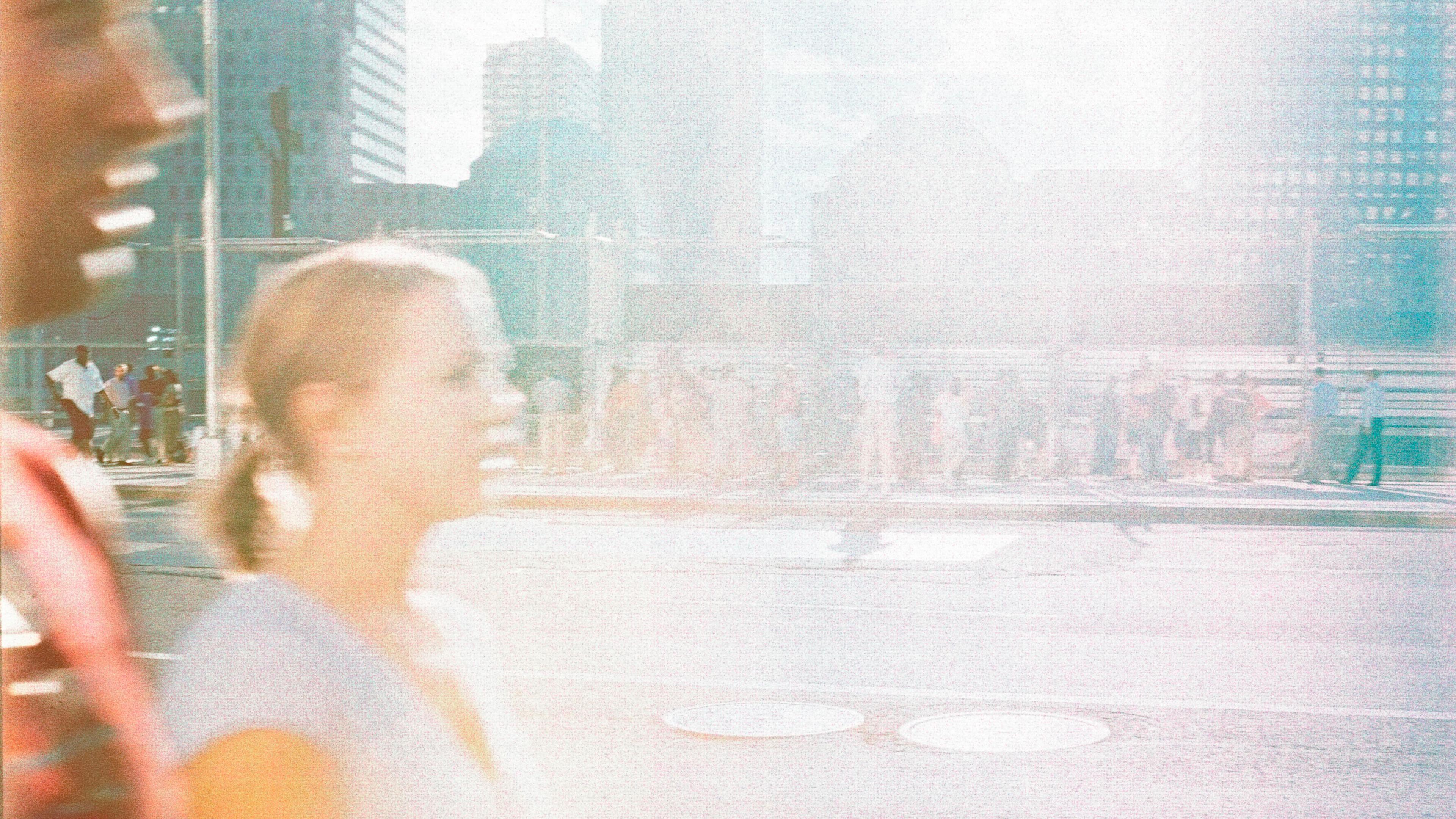 Photo of a city street with two blurry people walking in foreground bright light flaring over the scene in the afternoon.