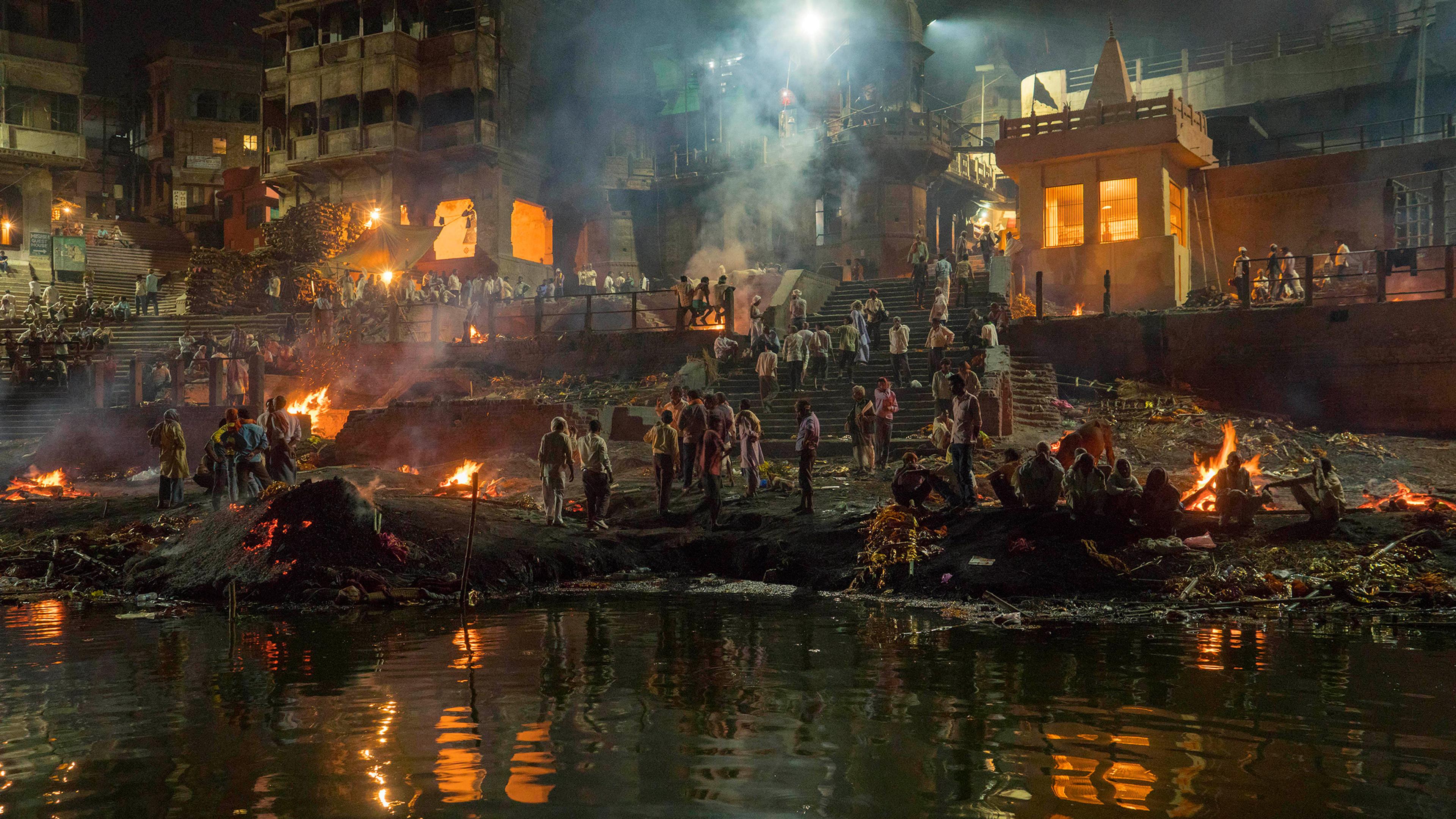 A cremation ceremony at night on a riverbank, surrounded by fires, people and historical buildings.