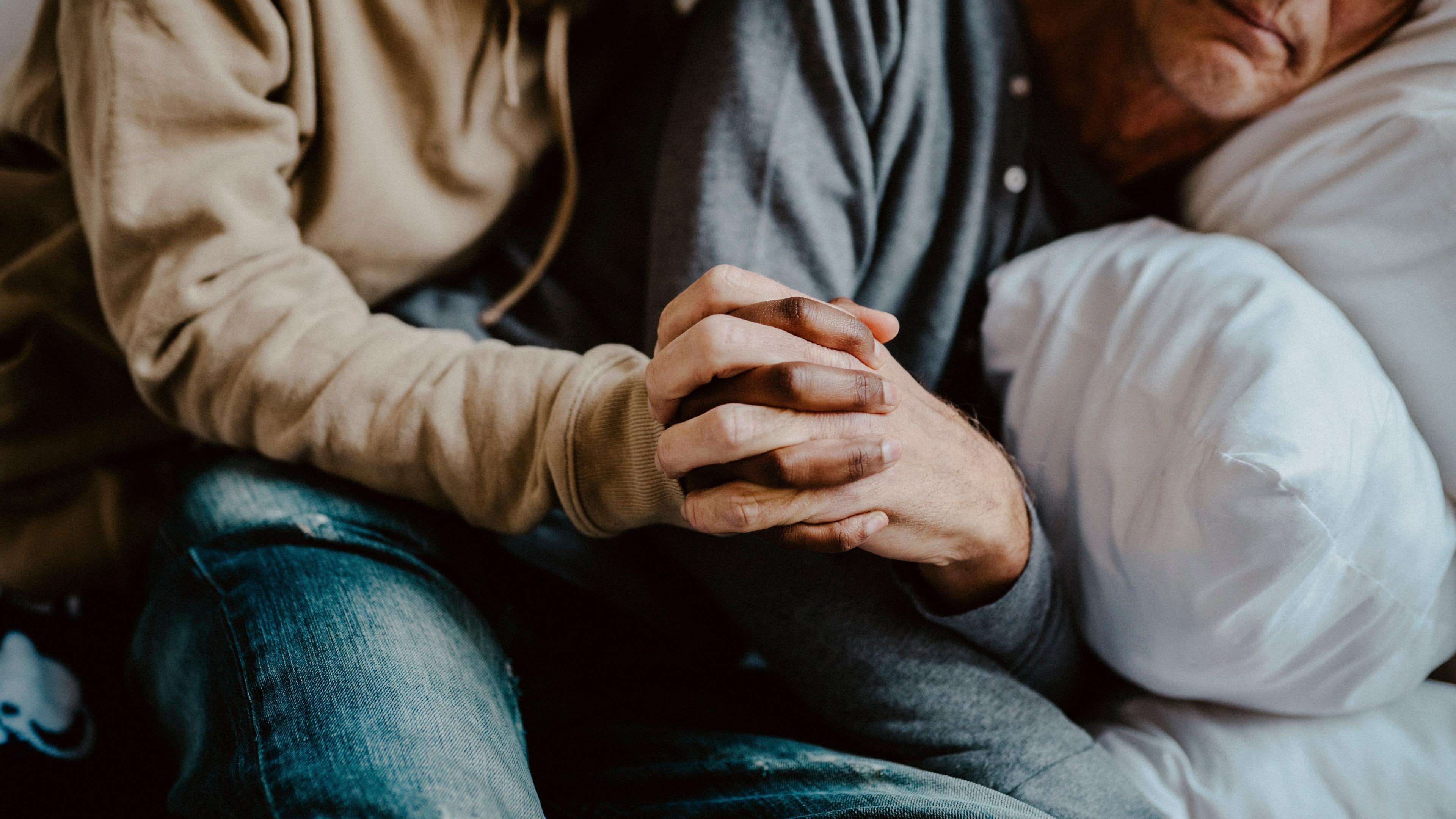 Photo of two people seated holding hands closely, one in a beige hoodie the other in a grey shirt resting on a pillow.