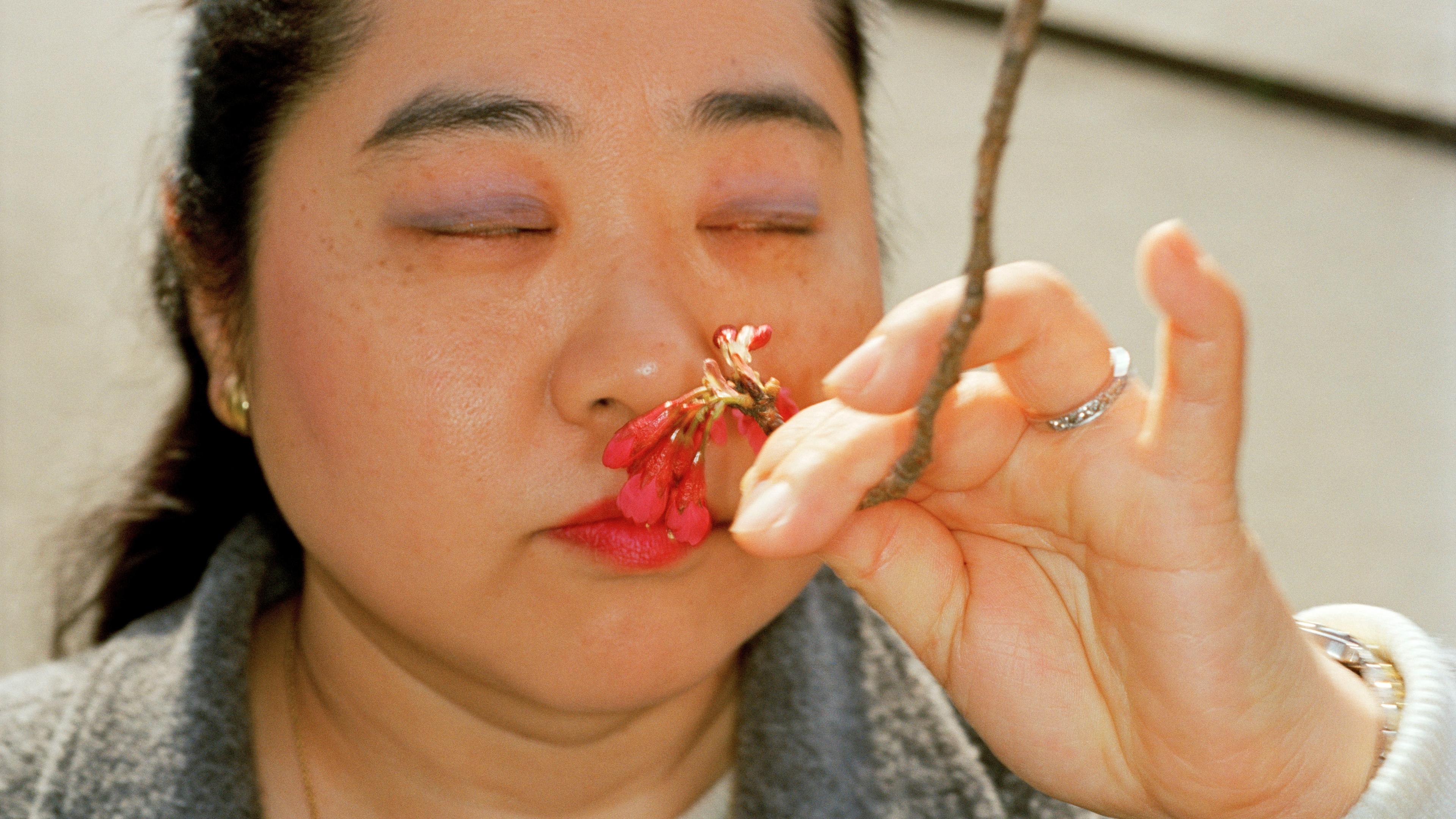 Close-up photo of a woman with closed eyes smelling a small branch with pink flowers held in her hand.