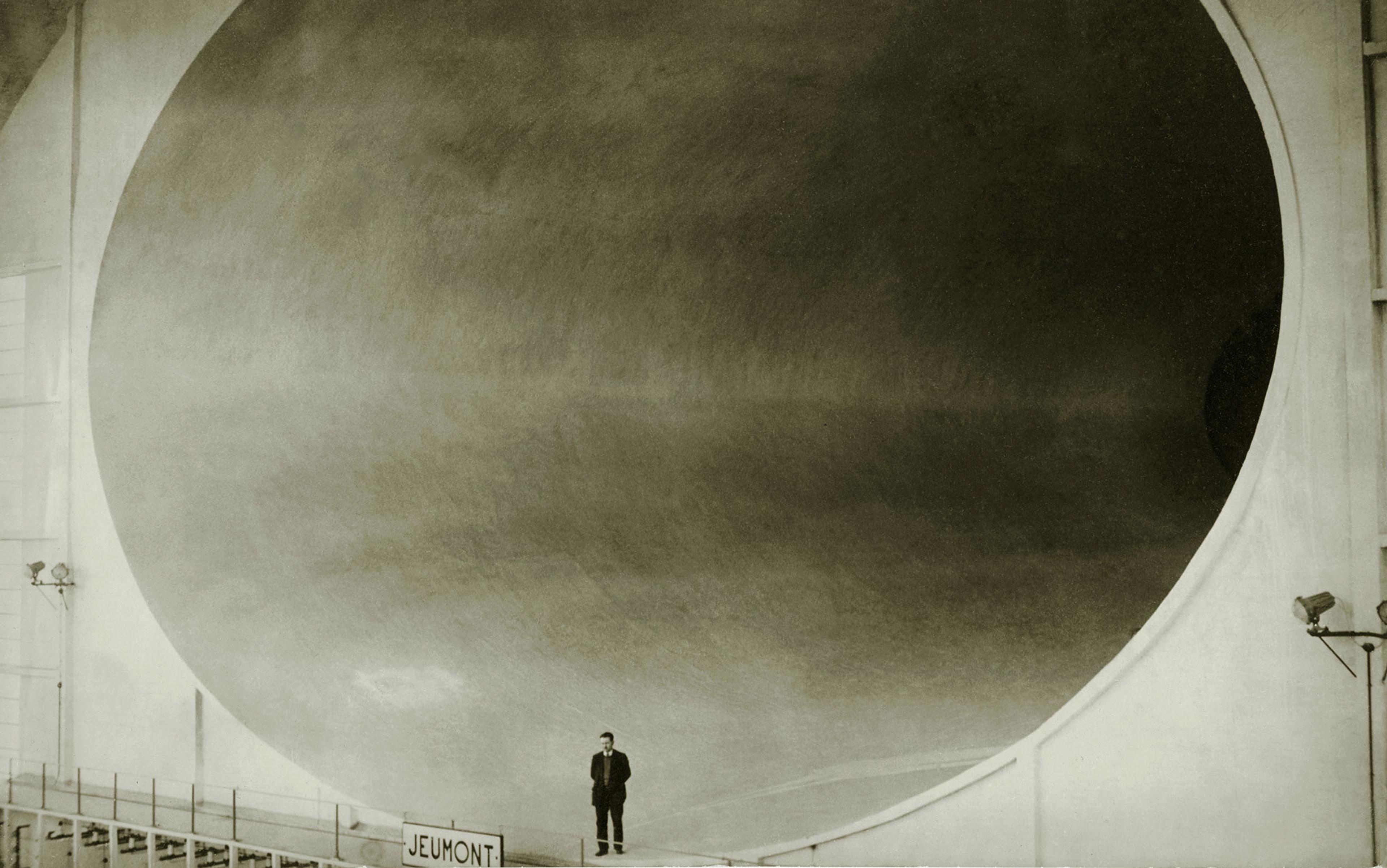 Black and white photo of man standing in front of a massive industrial tunnel with a Jeumont sign.