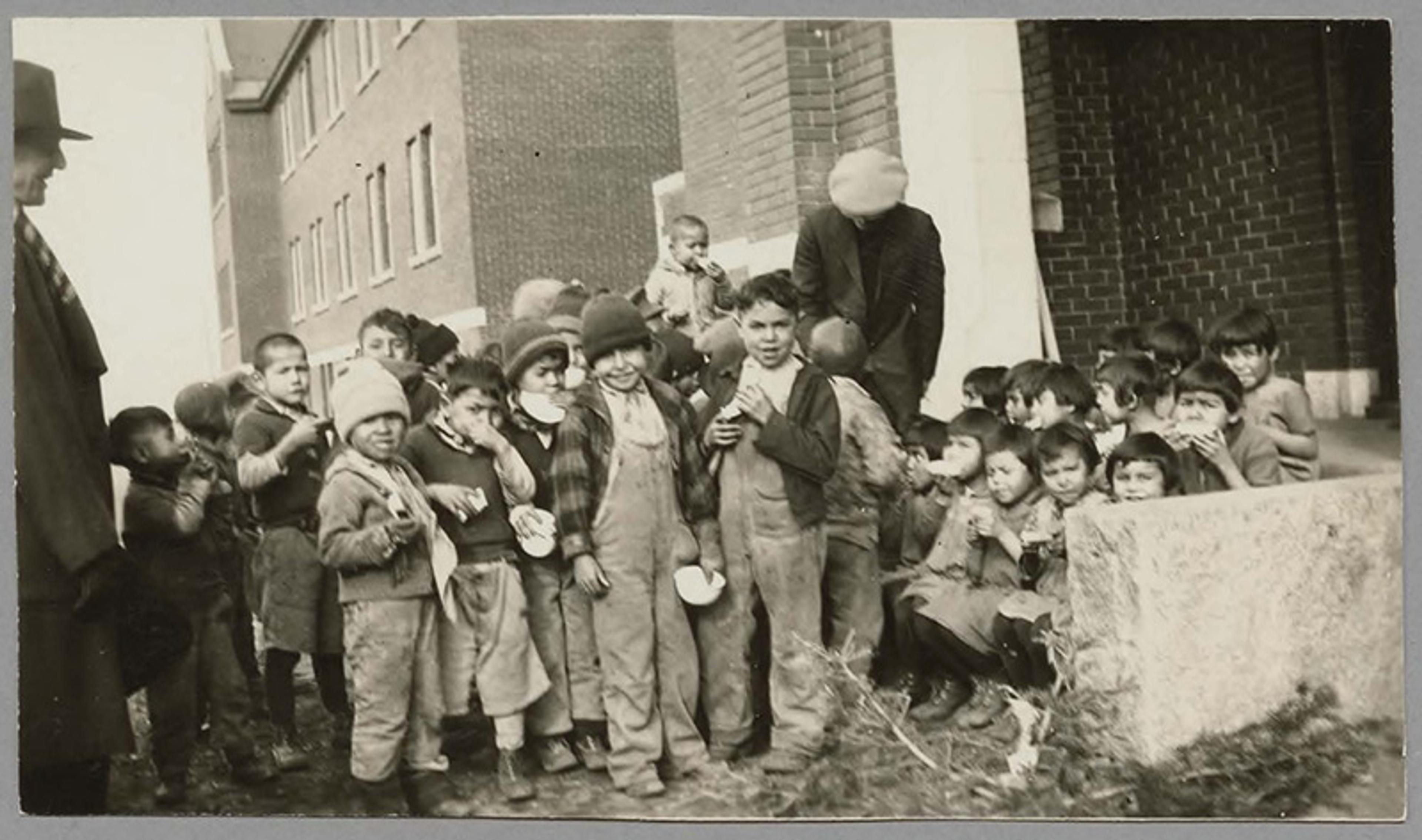 Black and white photo of children in winter clothing standing outside a brick building, likely in the early 20th century.