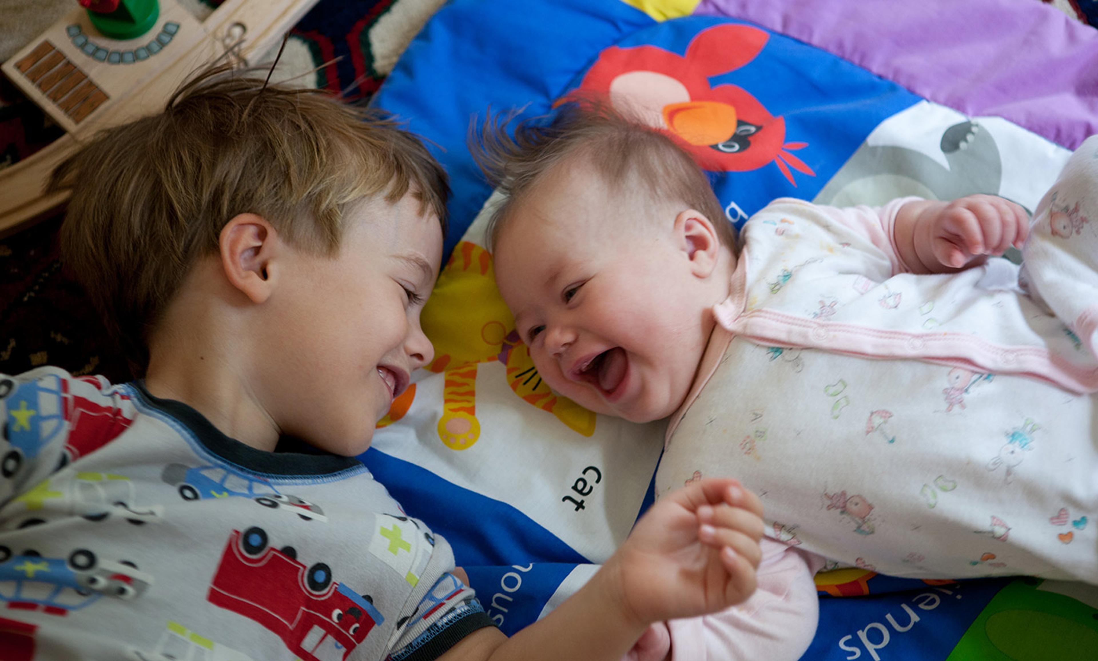 A smiling boy and a laughing baby lying down, facing each other on a colourful mat with toys around.