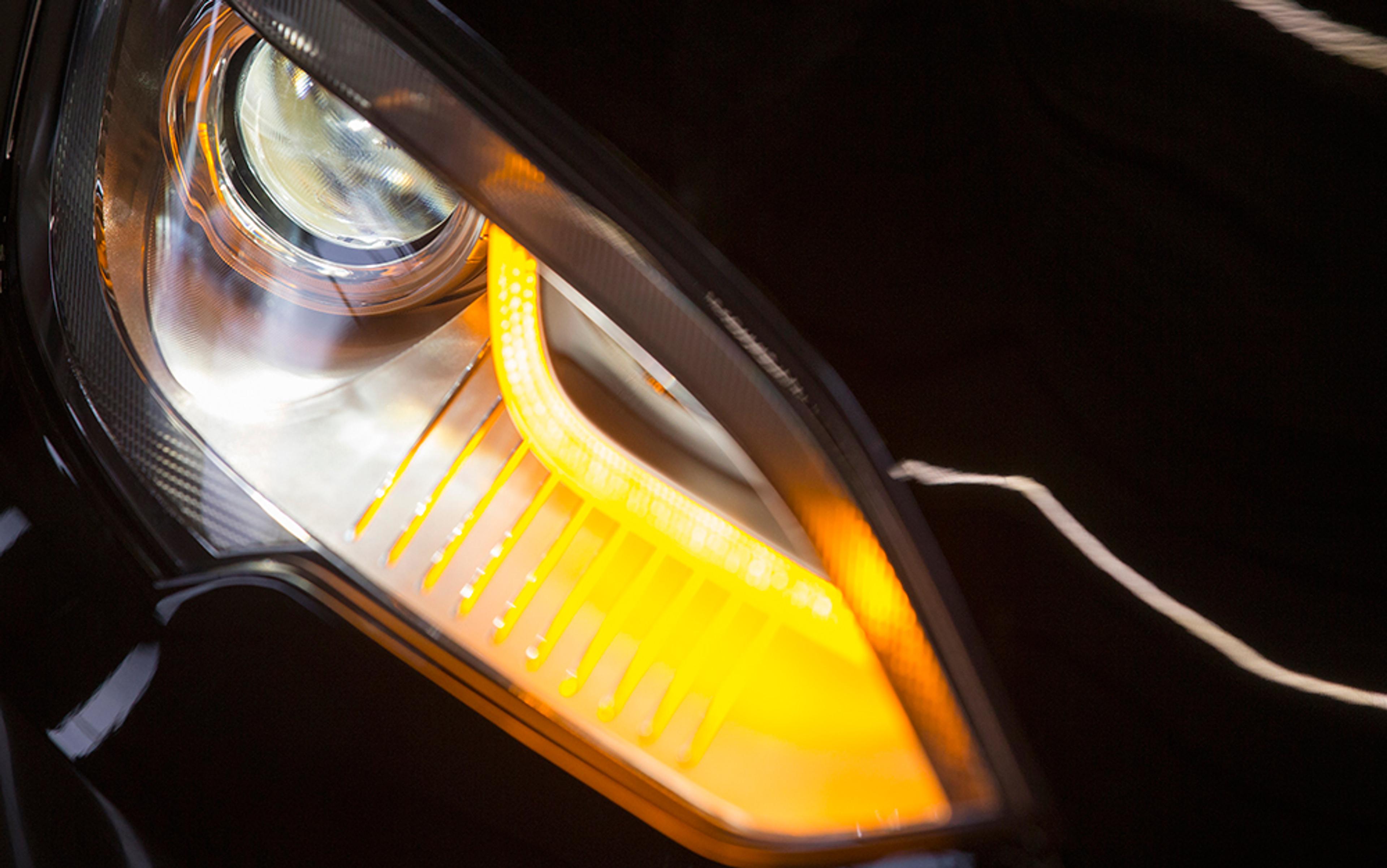 A close-up of a car headlight with illuminated yellow and white lights against a dark background.