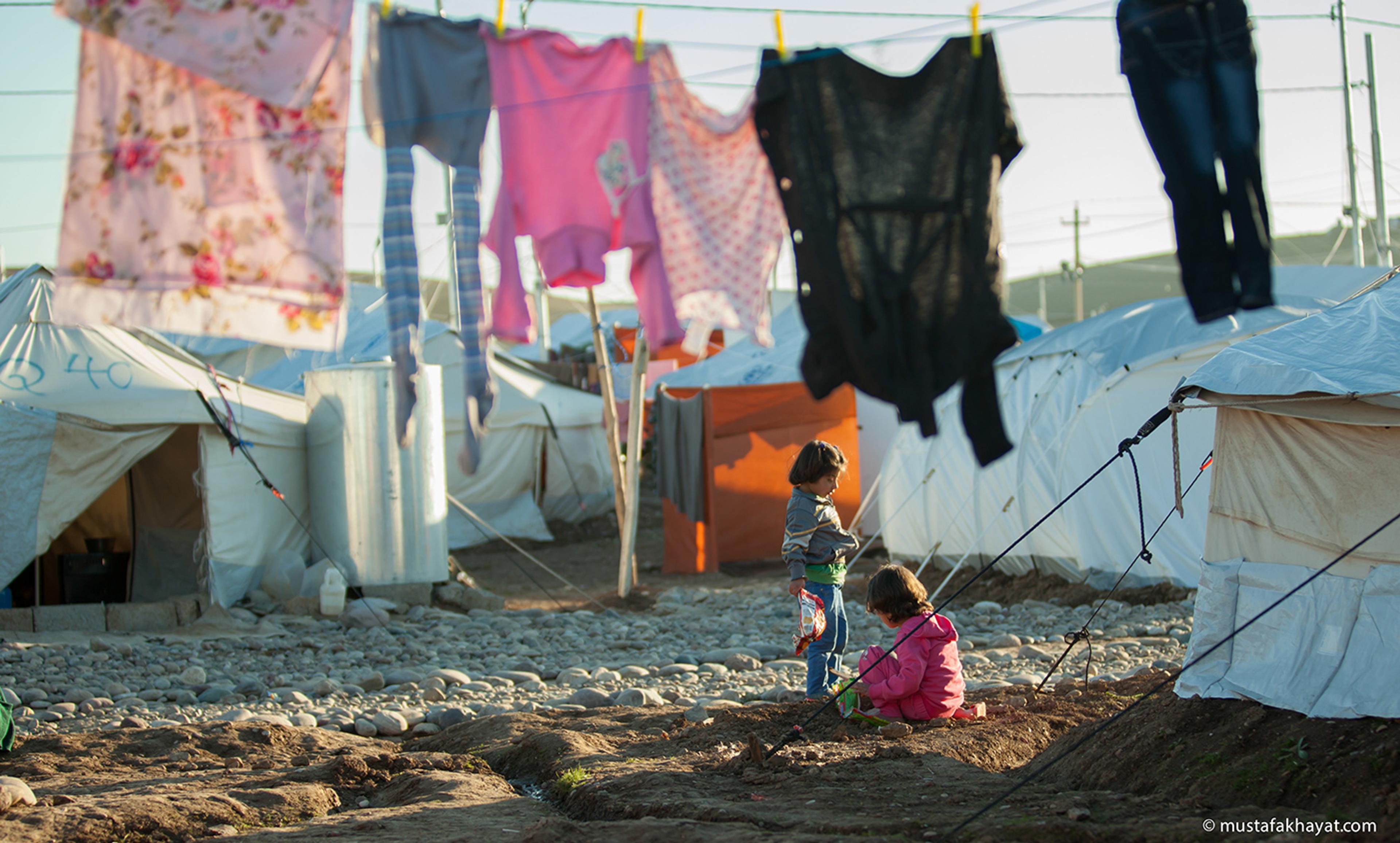Two children in a refugee camp with tents and clothes hanging on a line. The children are playing on a rocky dirt path.