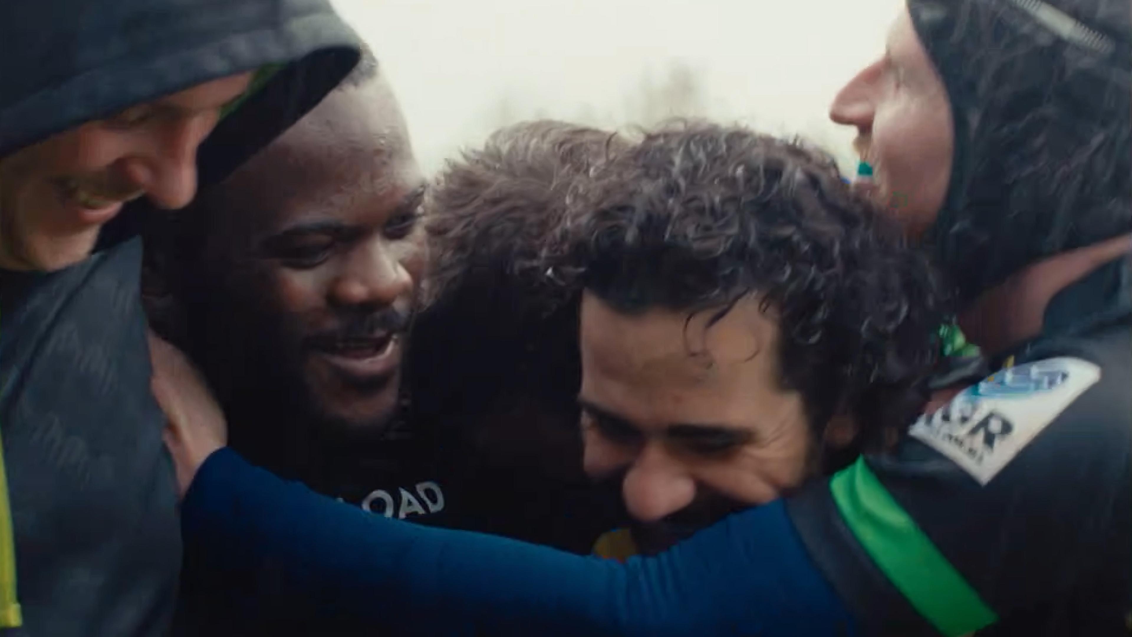 Photo of a group of rugby players wearing jerseys and helmets hugging and smiling after a match in a close-up shot.
