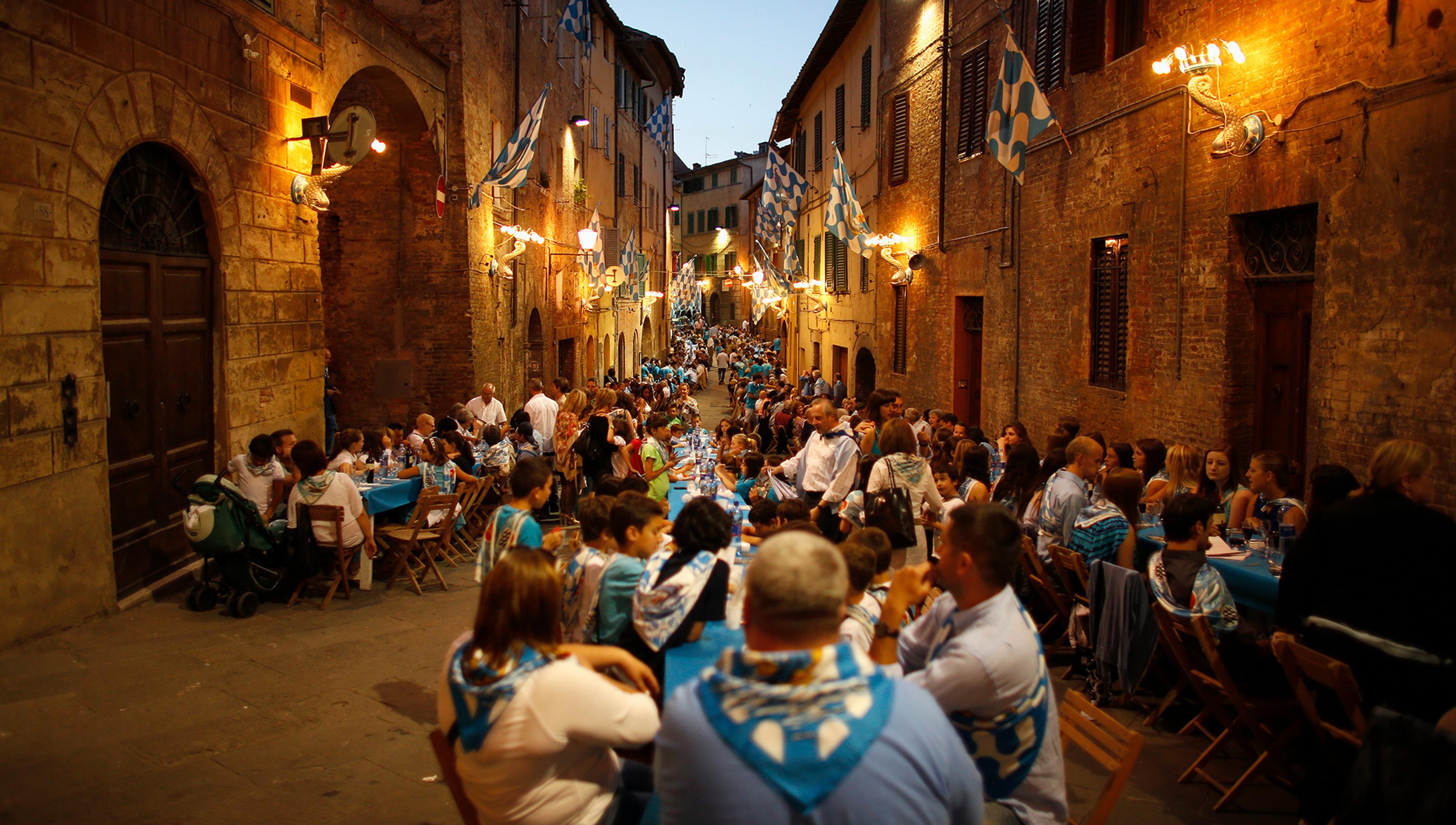 People dining at long tables adorned with blue tablecloths in a narrow, lantern-lit street lined with old buildings and blue-and-white flags.