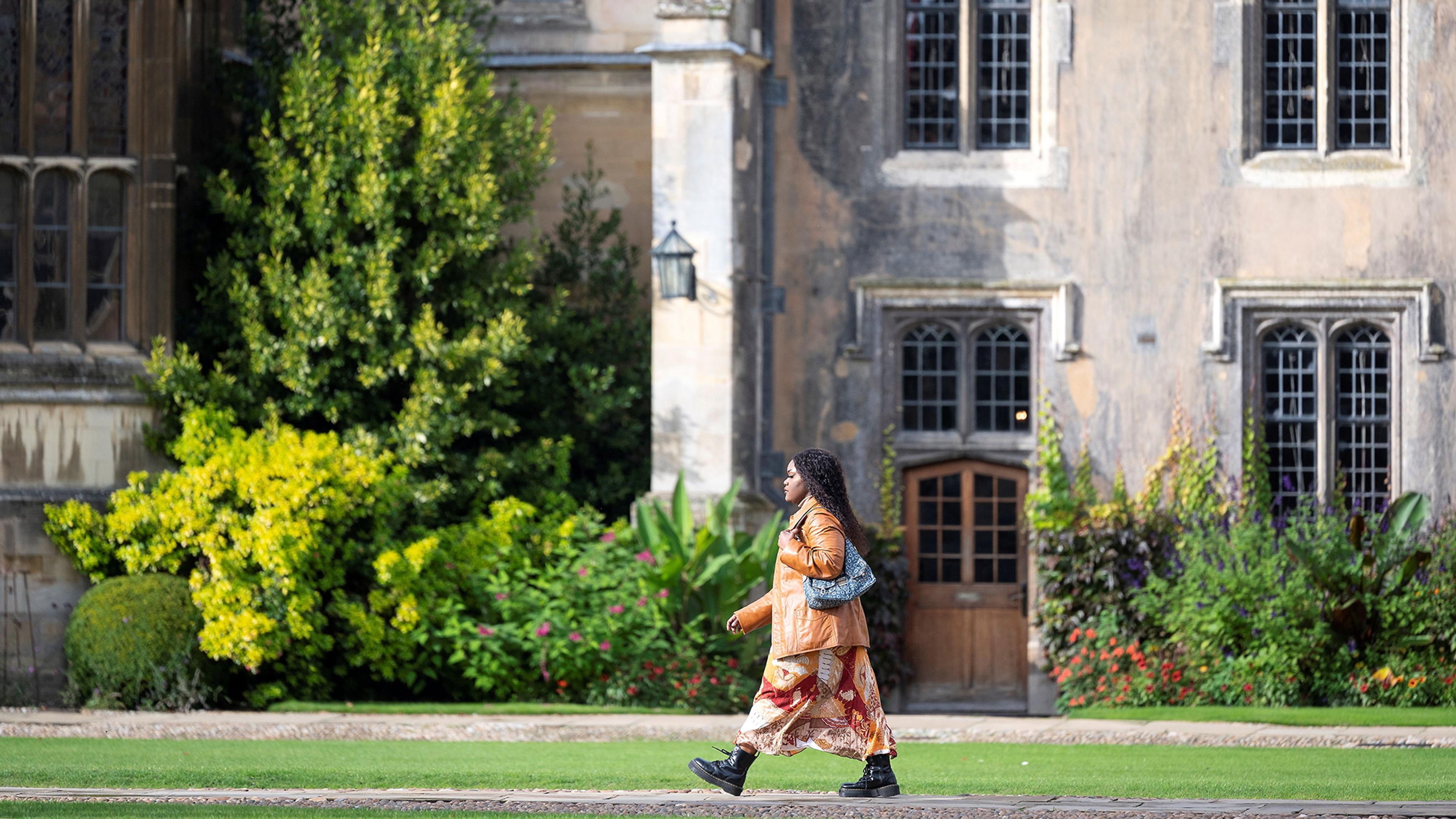 Photo of a person walking past a historic building with greenery and flowers in the background.