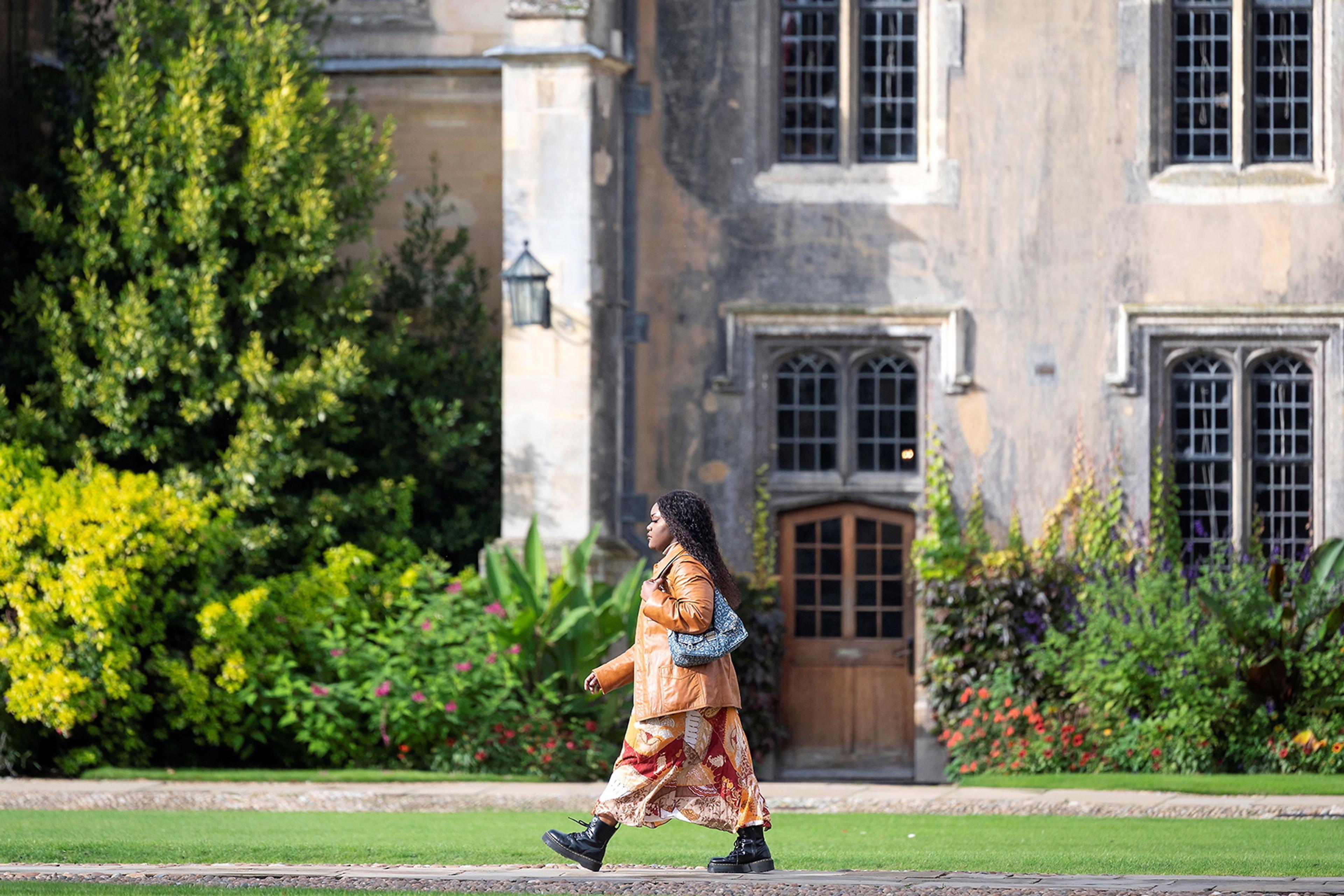 Photo of a person walking past a historic building with greenery and flowers in the background.