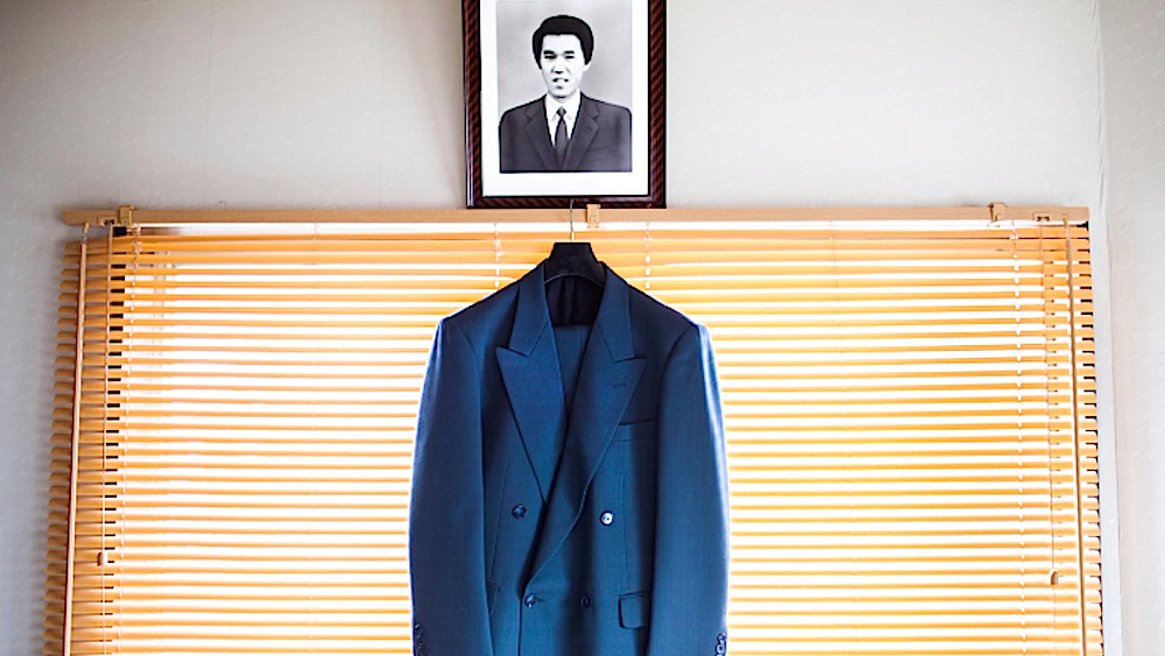 A blue suit jacket hanging on a coat hanger in front of closed blinds with a black and white portrait above it.