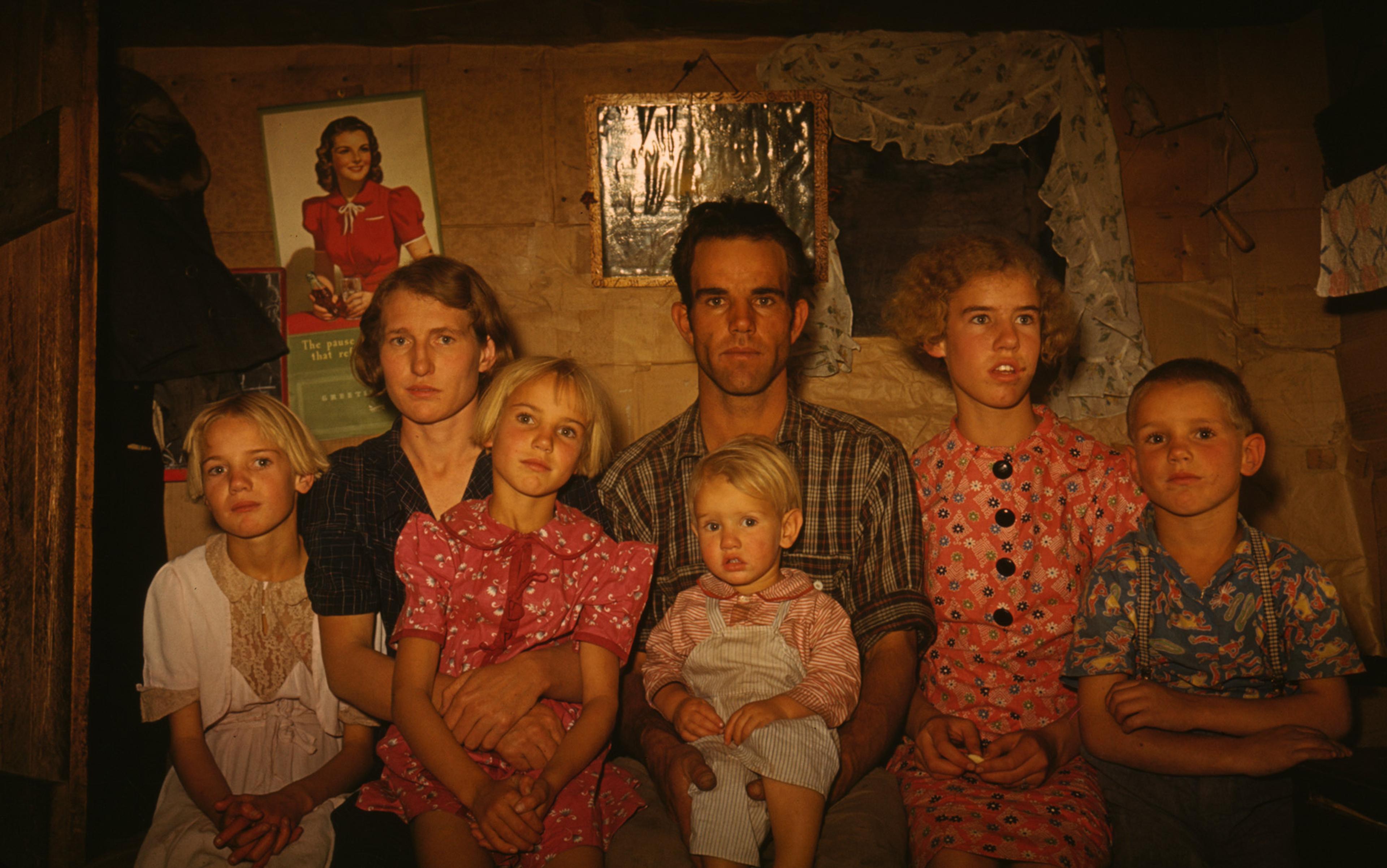 A family of with five children sitting closely together in a modest room with a pictures on the back wall.