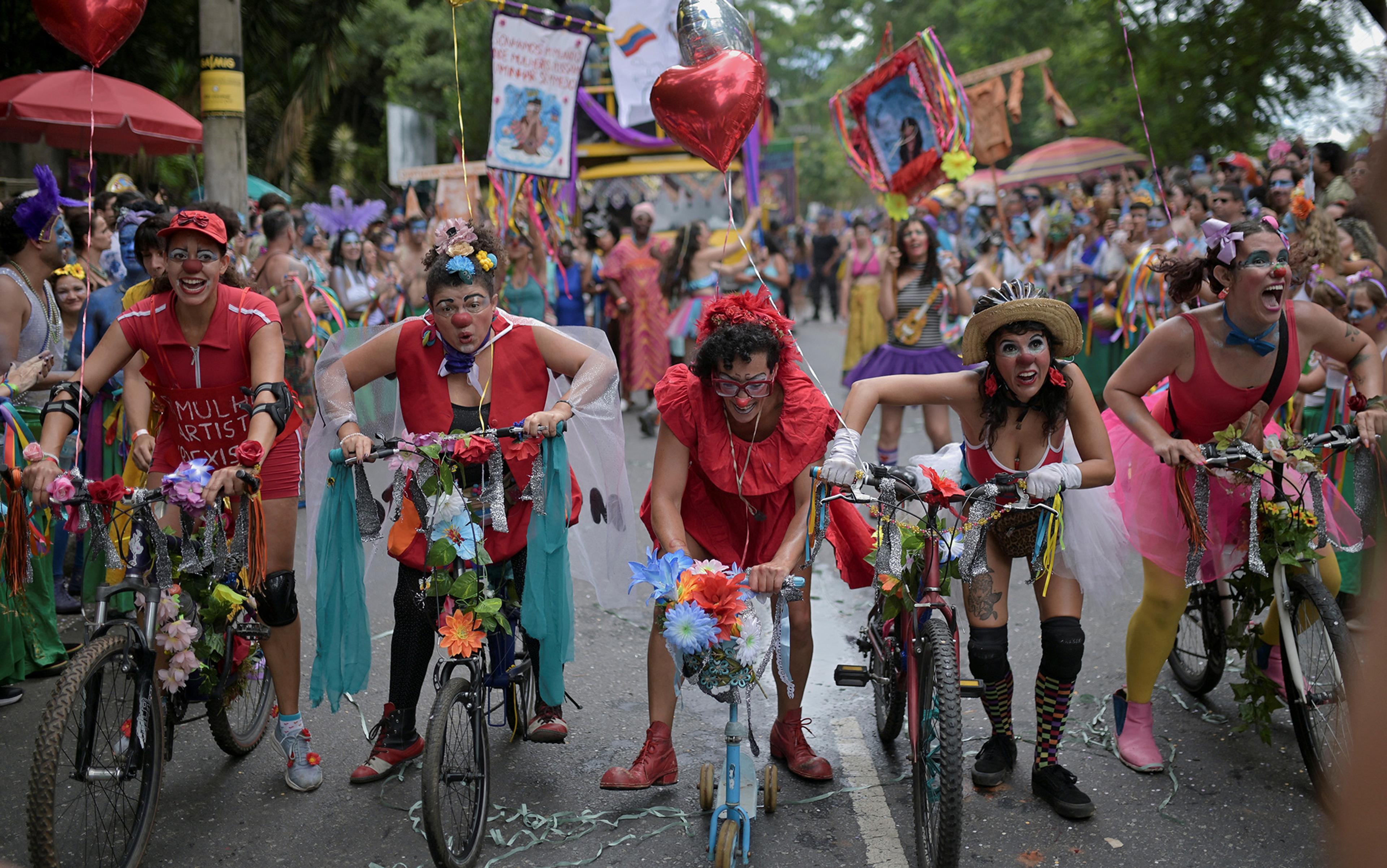 A vibrant parade with people dressed in colourful, clown-like costumes riding decorated bicycles. They are smiling and laughing as they ride through a crowd of onlookers. Various decorations such as flowers, balloons, and banners add to the festive atmosphere. Trees line the background of the street.