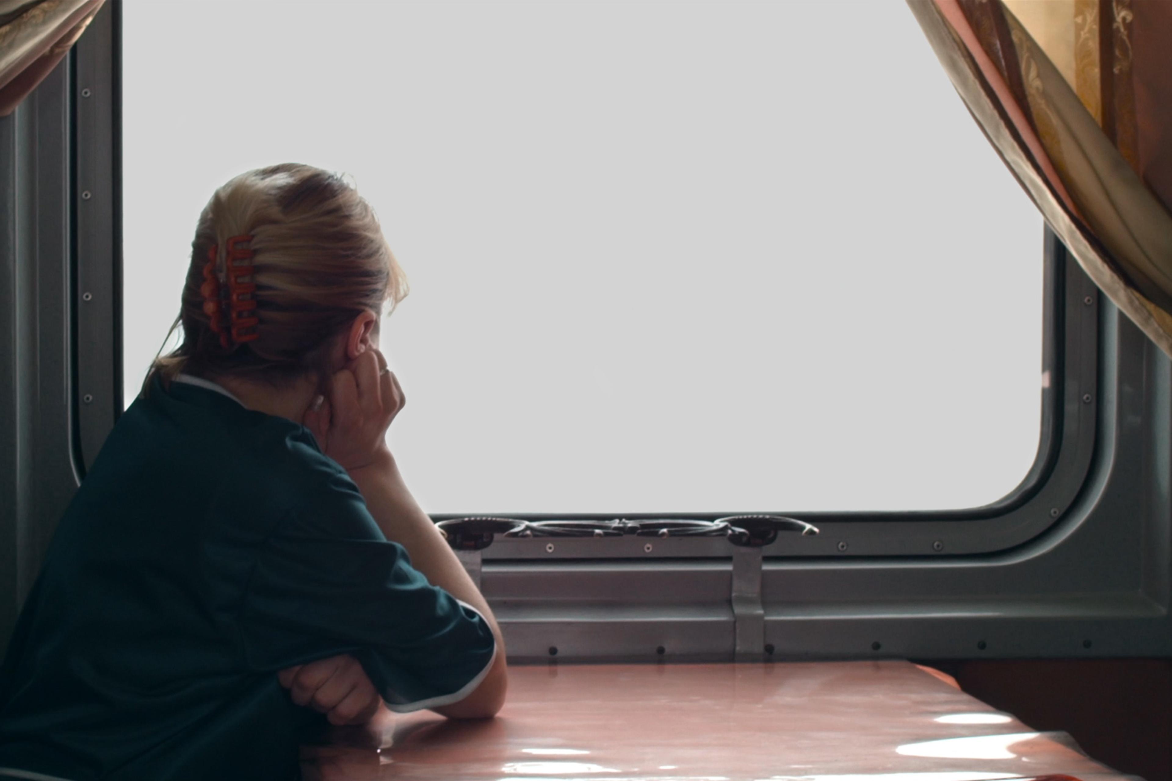 A woman with her hair up looking out of a train window, resting her chin on her hand. The outside view is white and indistinct.