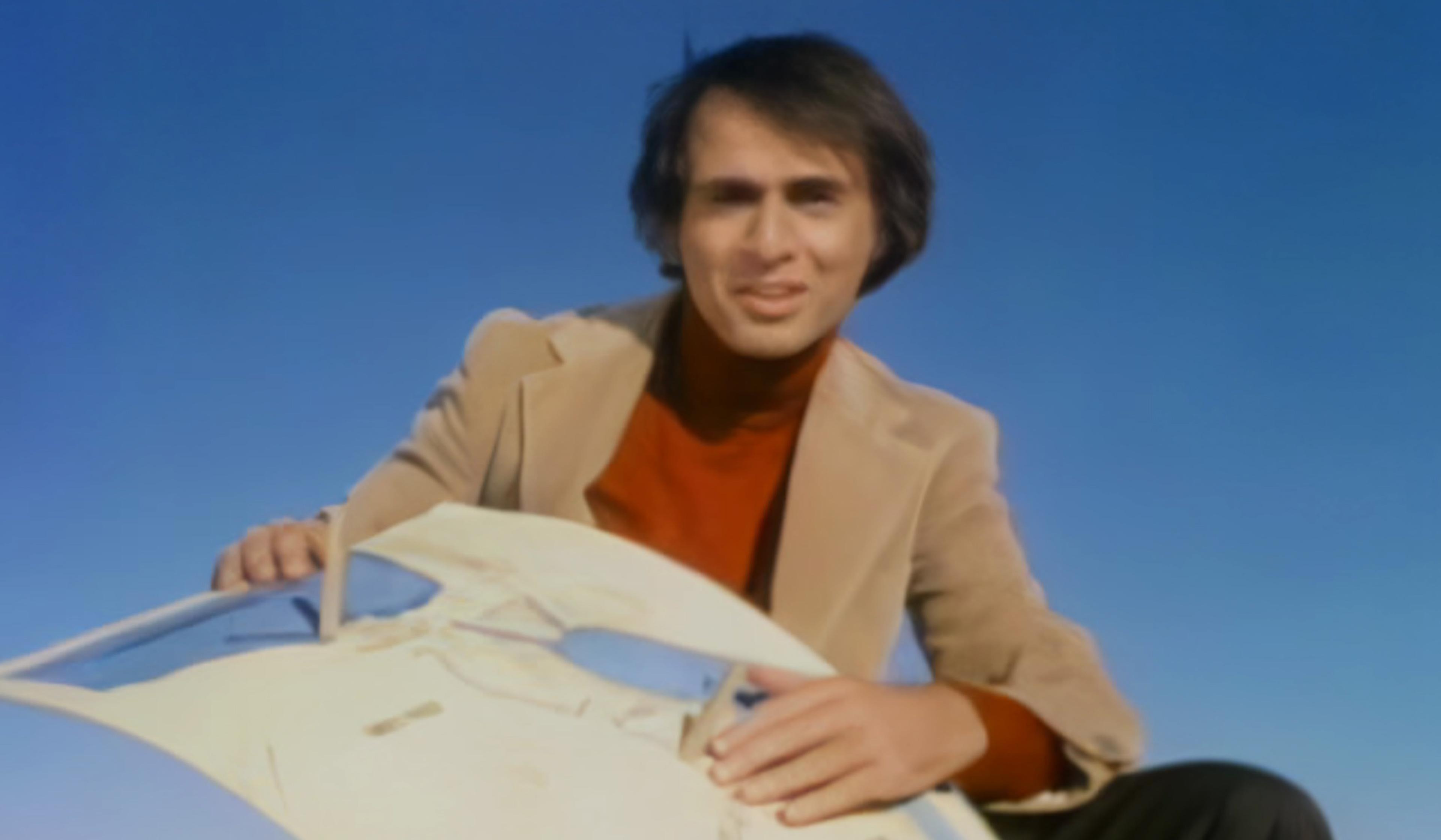 Carl Sagan in a beige blazer and red jumper holding a model against a clear blue sky.