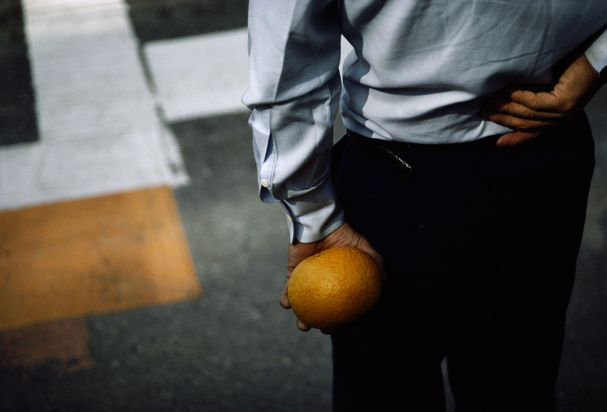 Photo of a person in a white shirt holding an orange behind their back at a crosswalk, seen from behind.