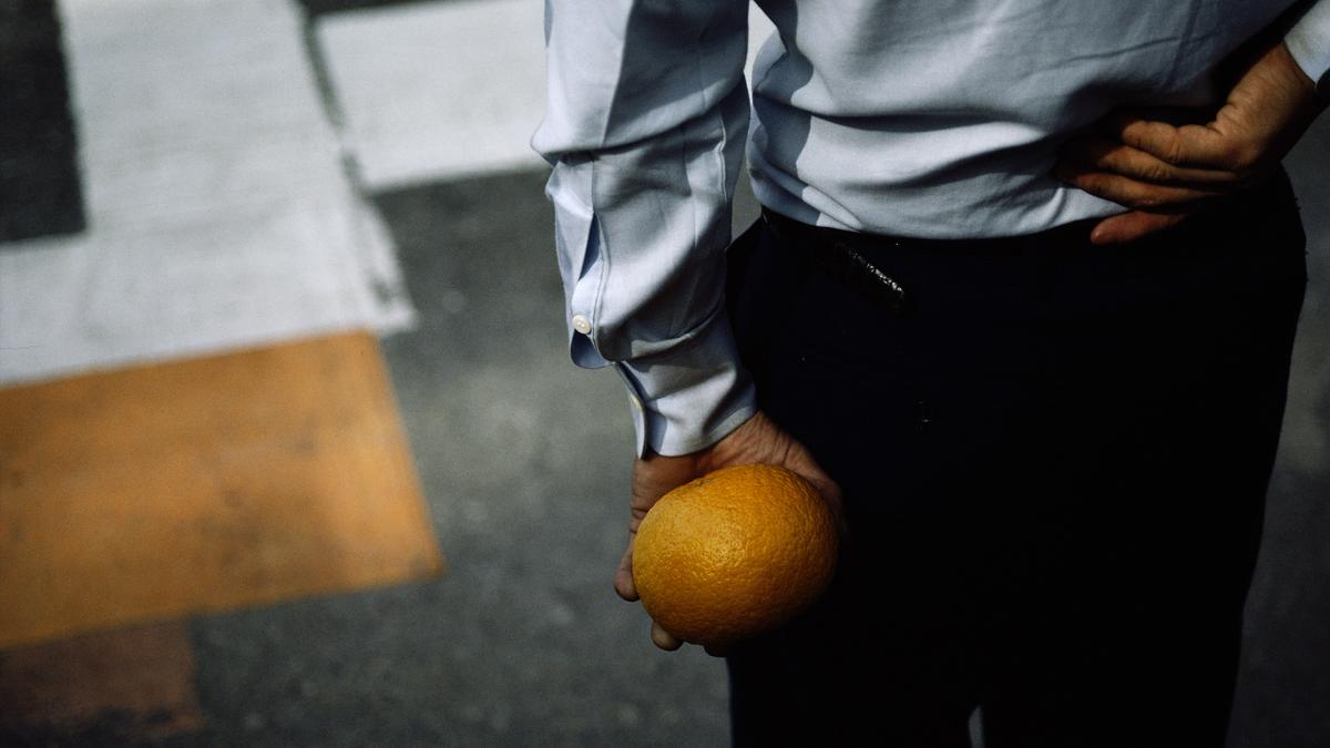 Photo of a person in a white shirt holding an orange behind their back at a crosswalk, seen from behind.