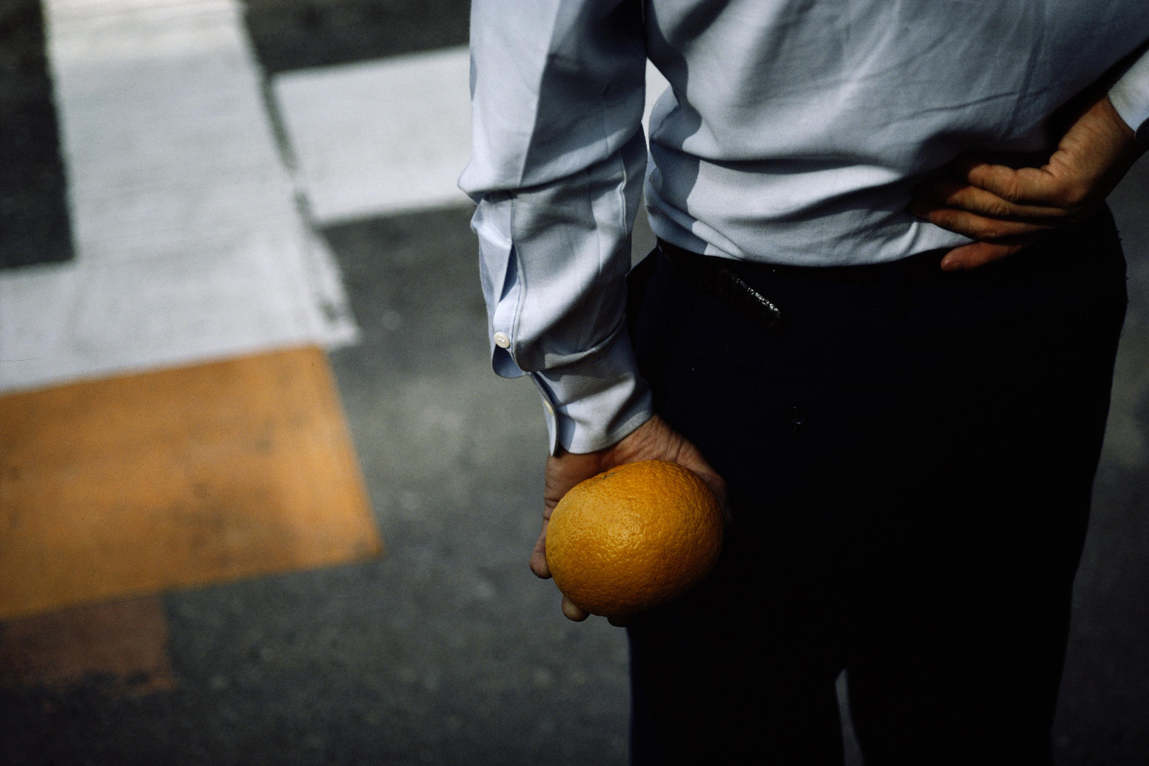 Photo of a person in a white shirt holding an orange behind their back at a crosswalk, seen from behind.
