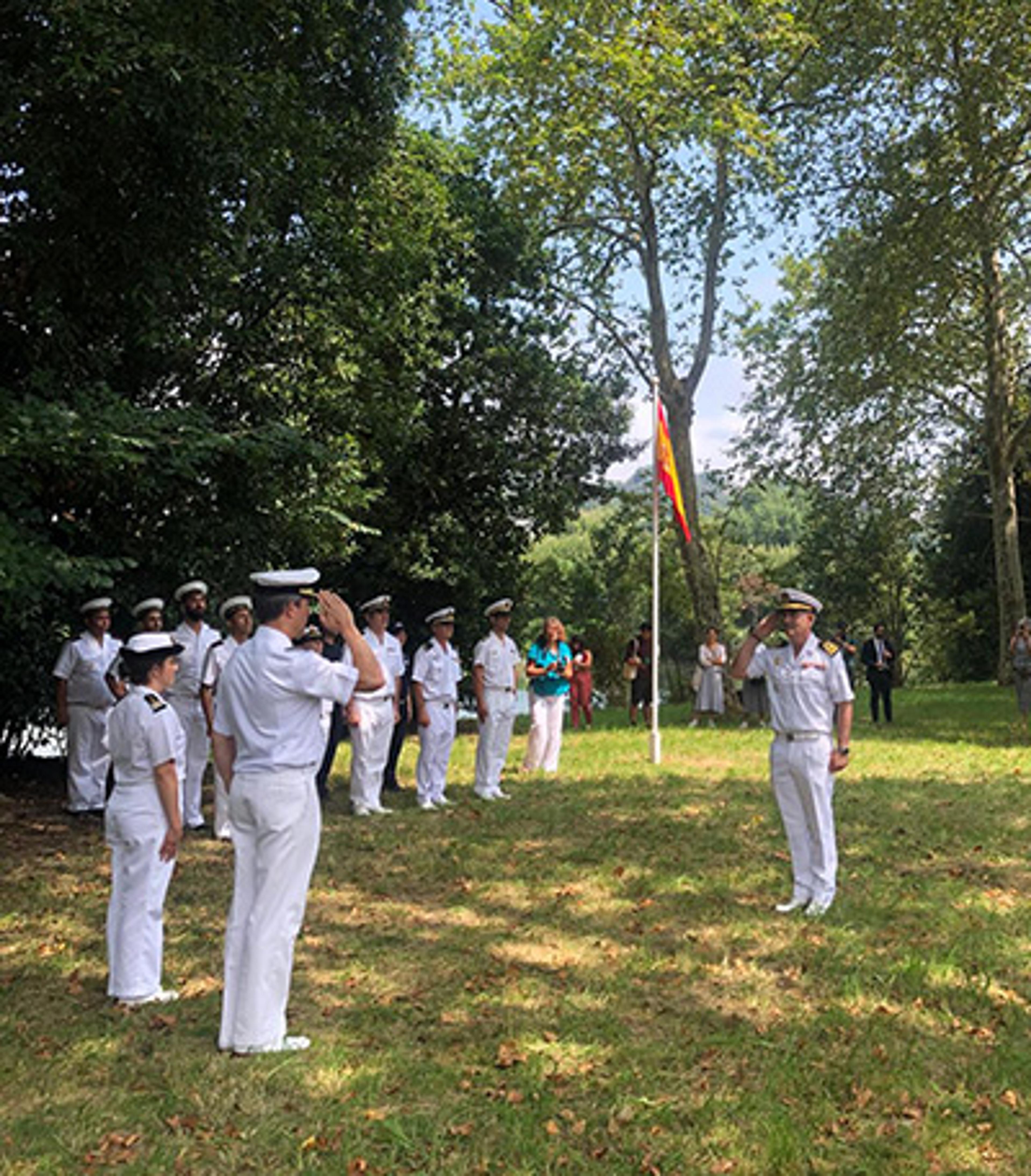 Photo of naval officers in white uniforms saluting near a flag on a grassy area with trees in the background.