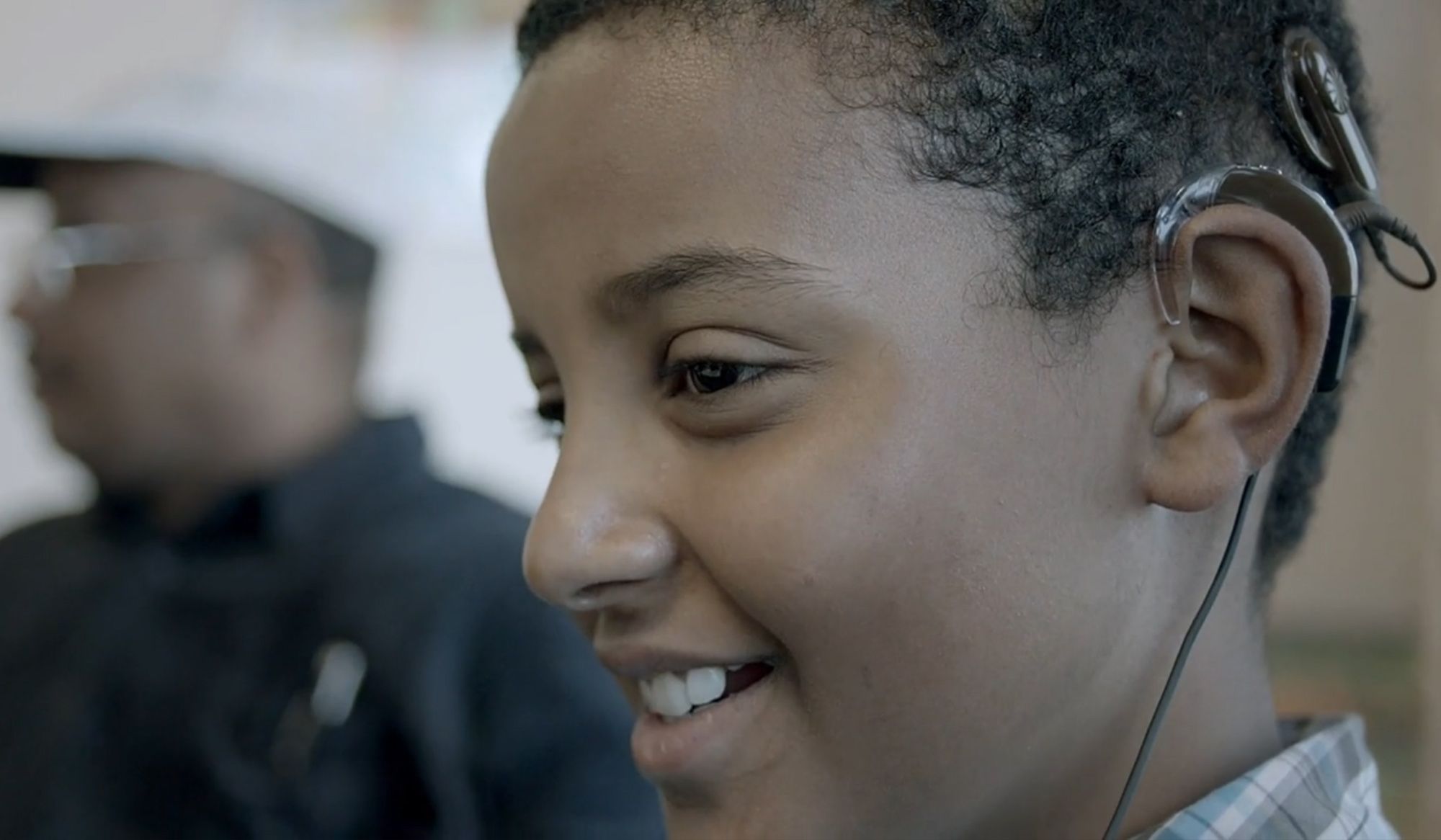 Close-up of a smiling boy wearing a hearing aid, with an out-of-focus adult in the background.