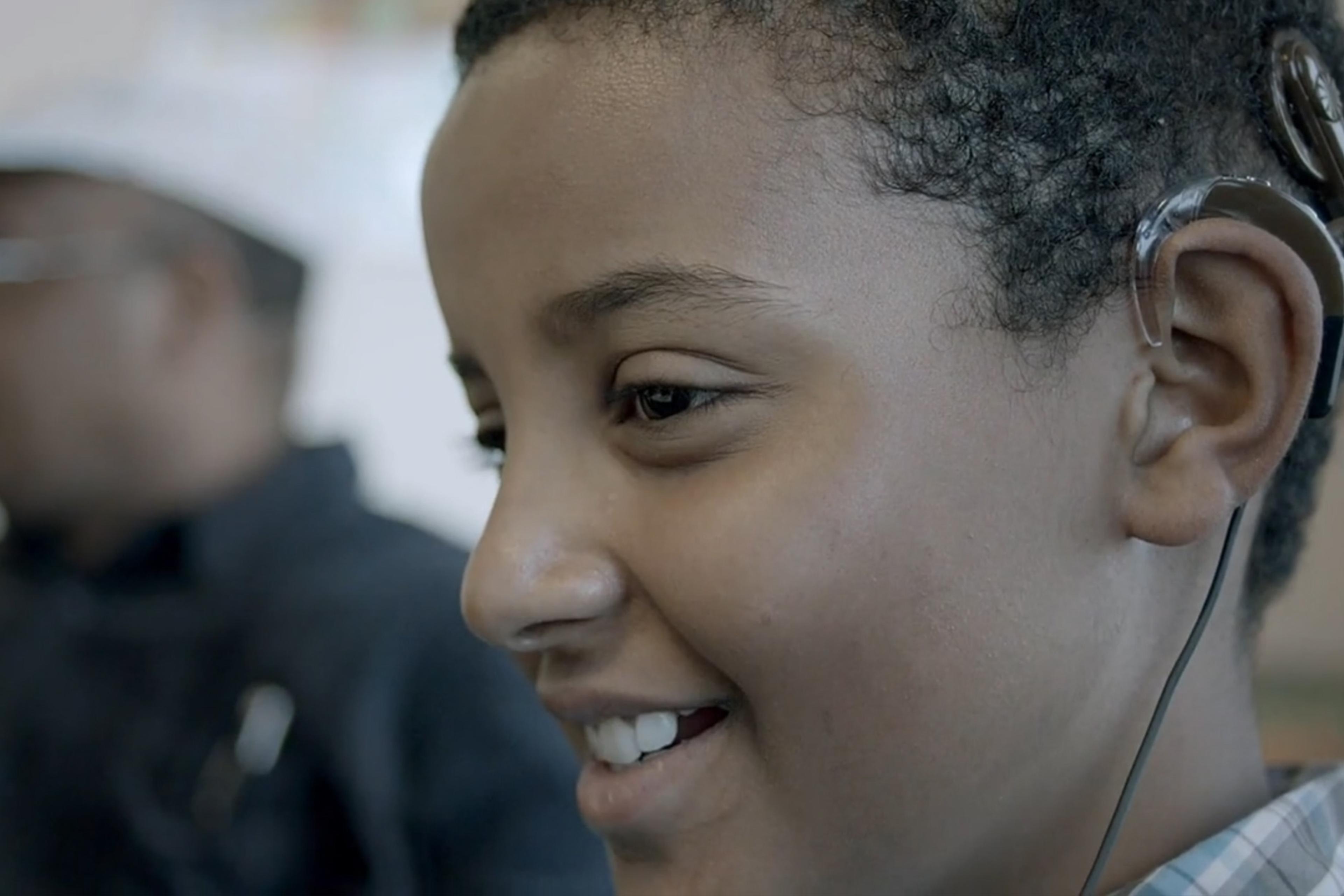 Close-up of a smiling boy wearing a hearing aid, with an out-of-focus adult in the background.
