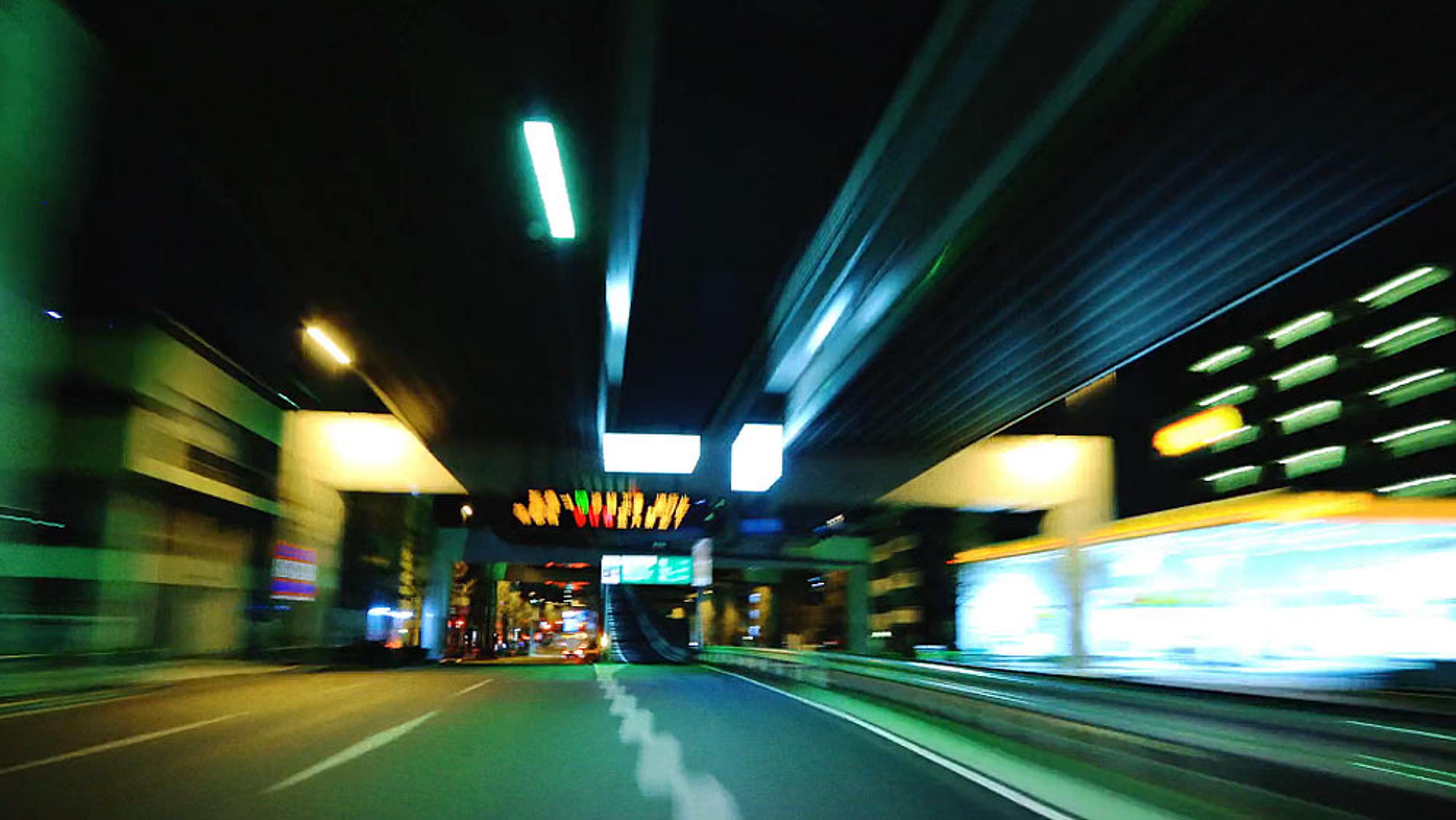 Blurred night photo of an urban road with bright lights, underpasses, and buildings, conveying a sense of speed and motion.