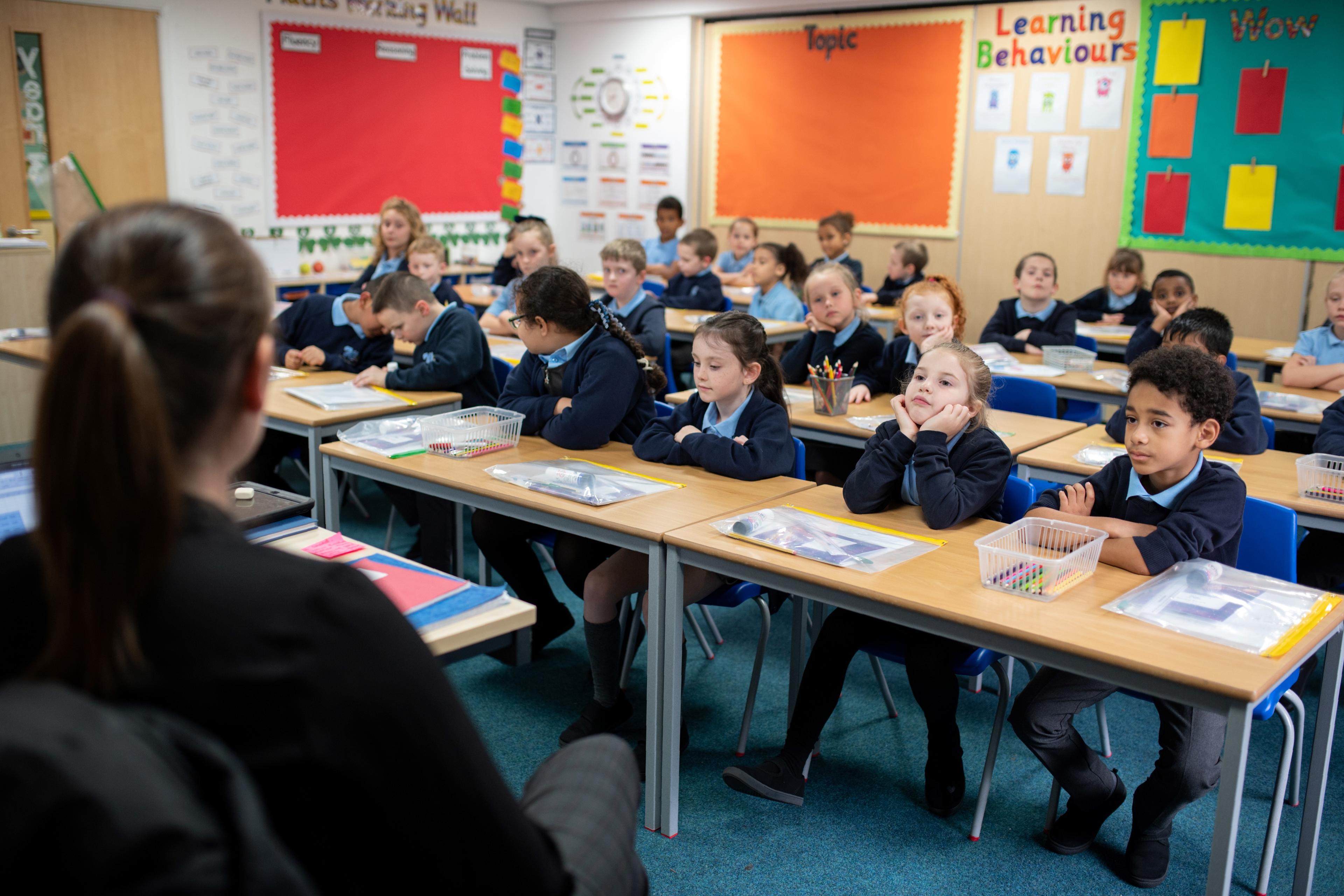 Photo of a classroom with young students seated at desks, facing a teacher. Colourful educational posters on the walls.
