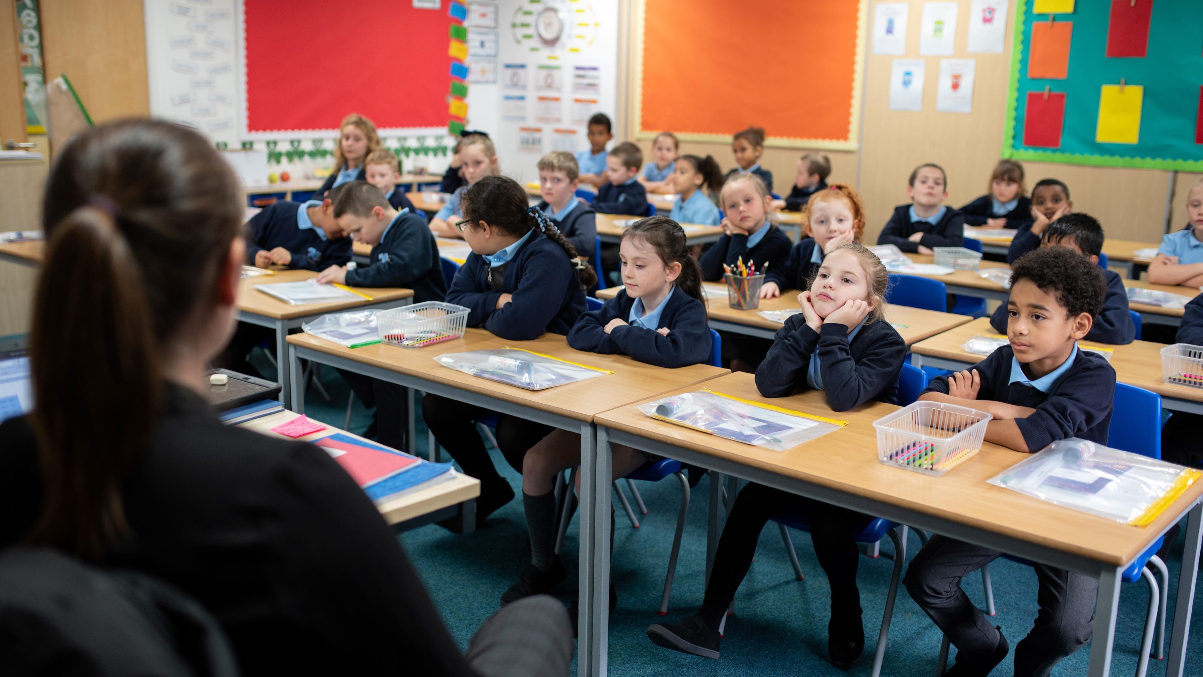 Photo of a classroom with young students seated at desks, facing a teacher. Colourful educational posters on the walls.