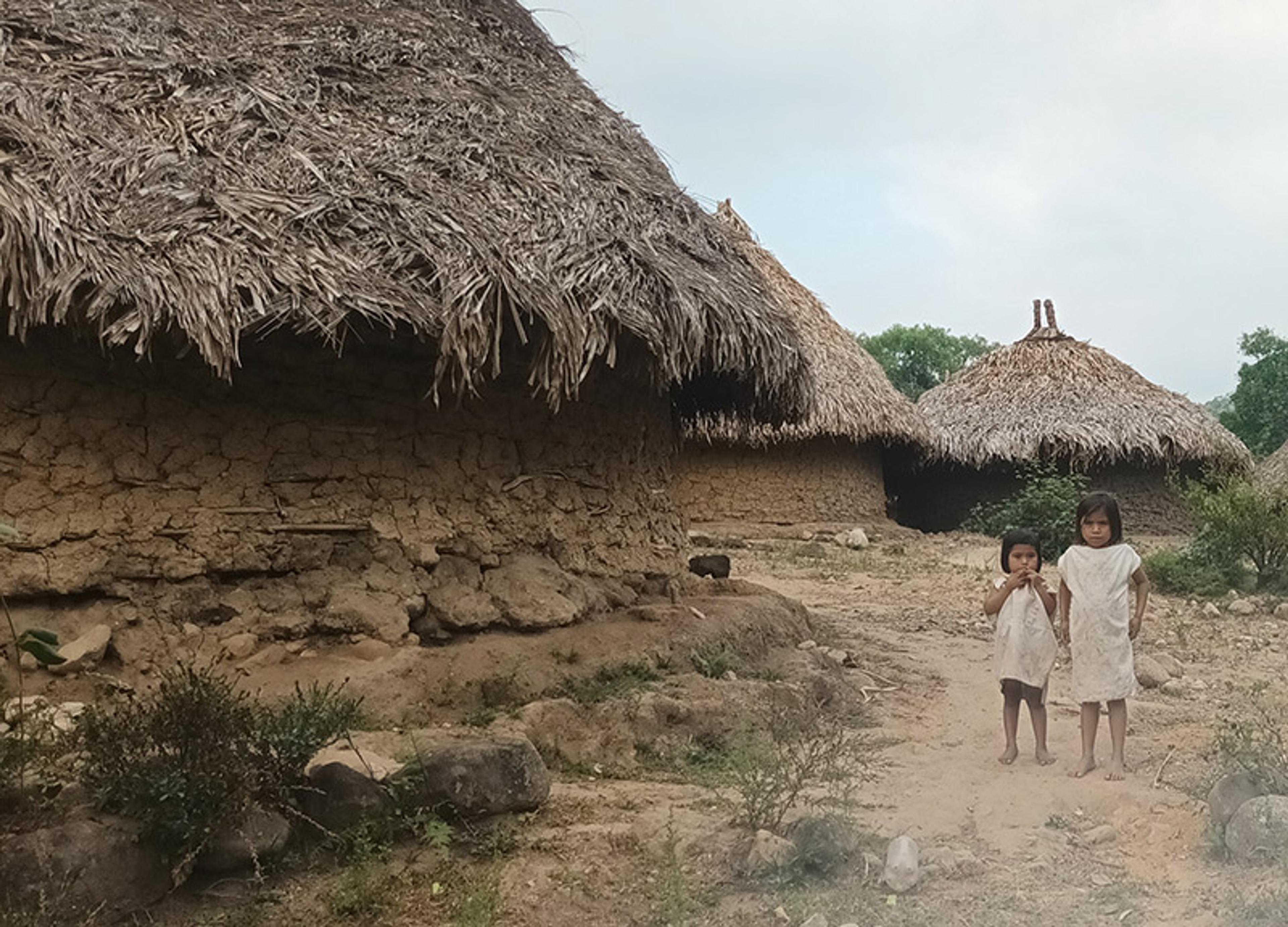 Two Colombian children standing in front of thatched mud huts in a rural setting with sparse vegetation and a cloudy sky.