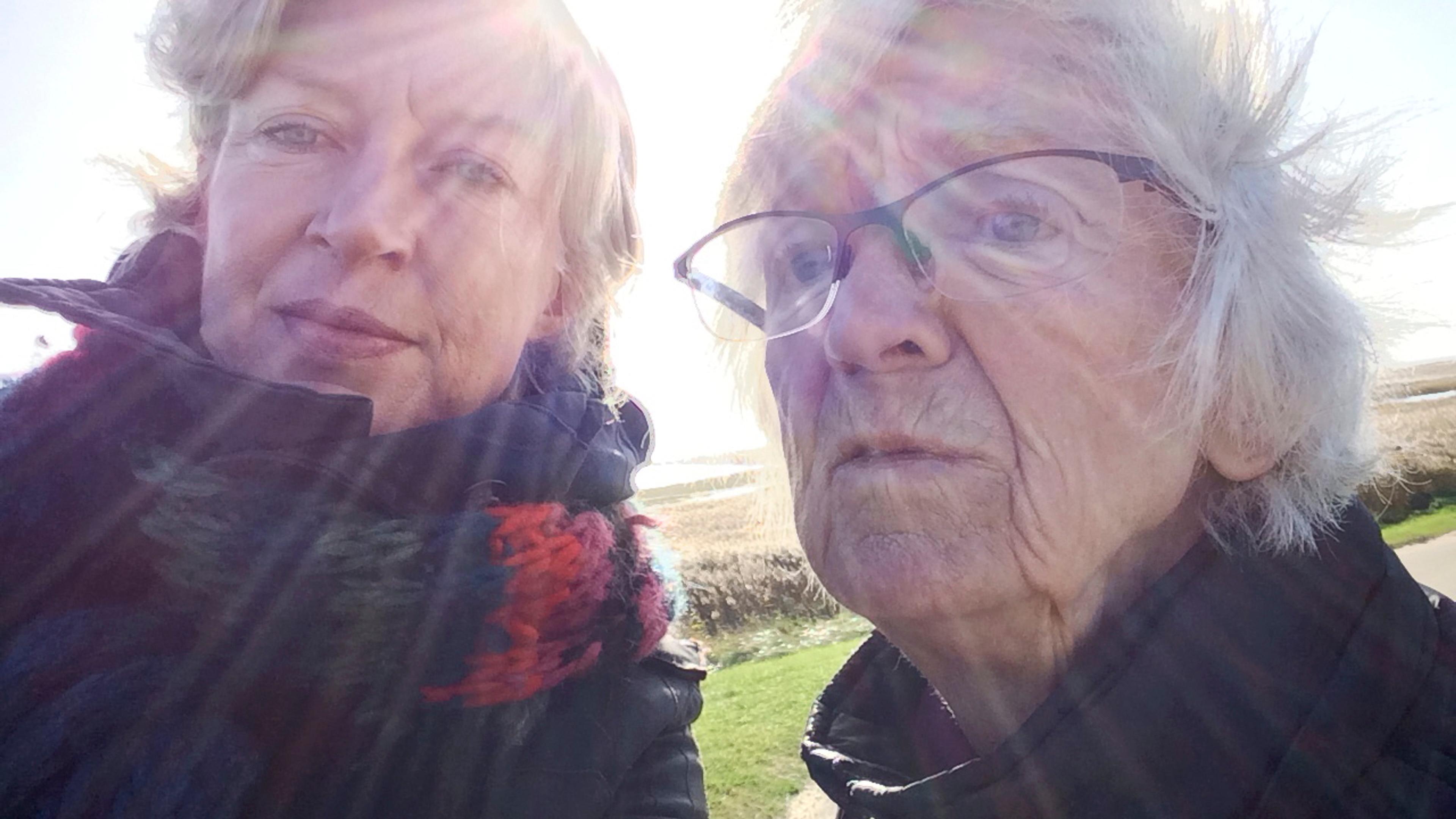 Photo of two people outdoors in winter clothing with sun rays surrounding them. One person wears glasses. Bright sky in background.