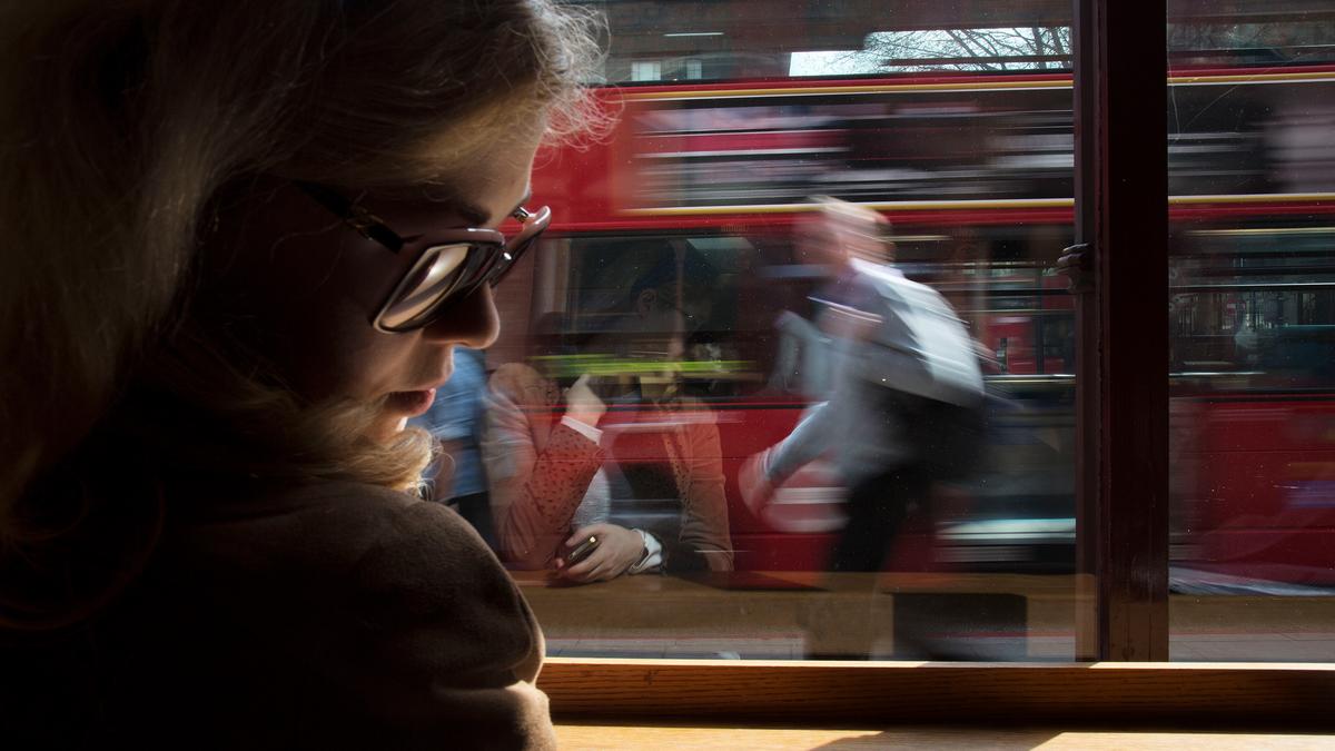 A woman inside wearing sunglasses, with a blurred reflection of people and a red bus moving outside the window.