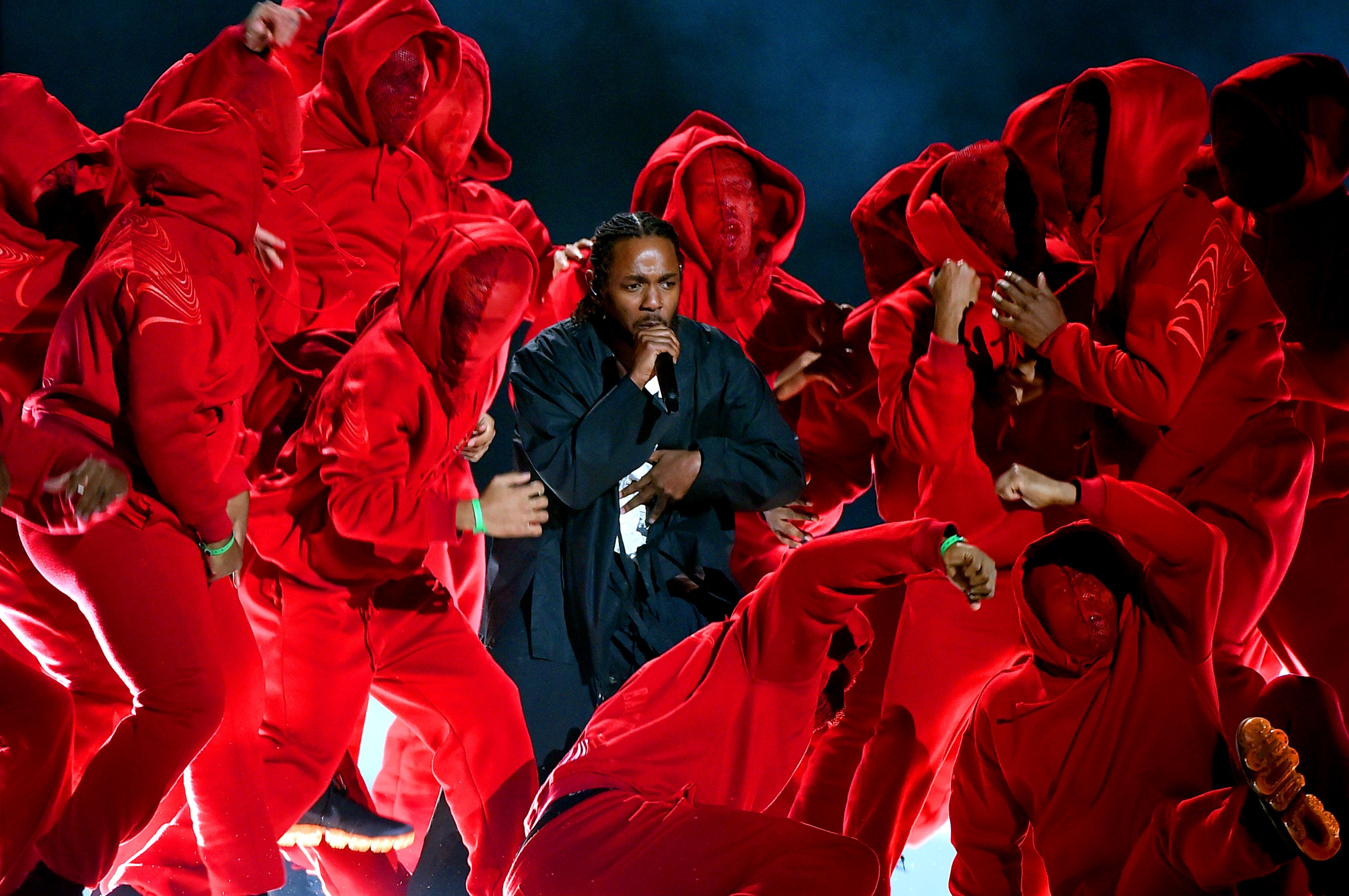 Photo of a performer singing surrounded by a group of dancers in red hooded outfits on stage under dramatic lighting.
