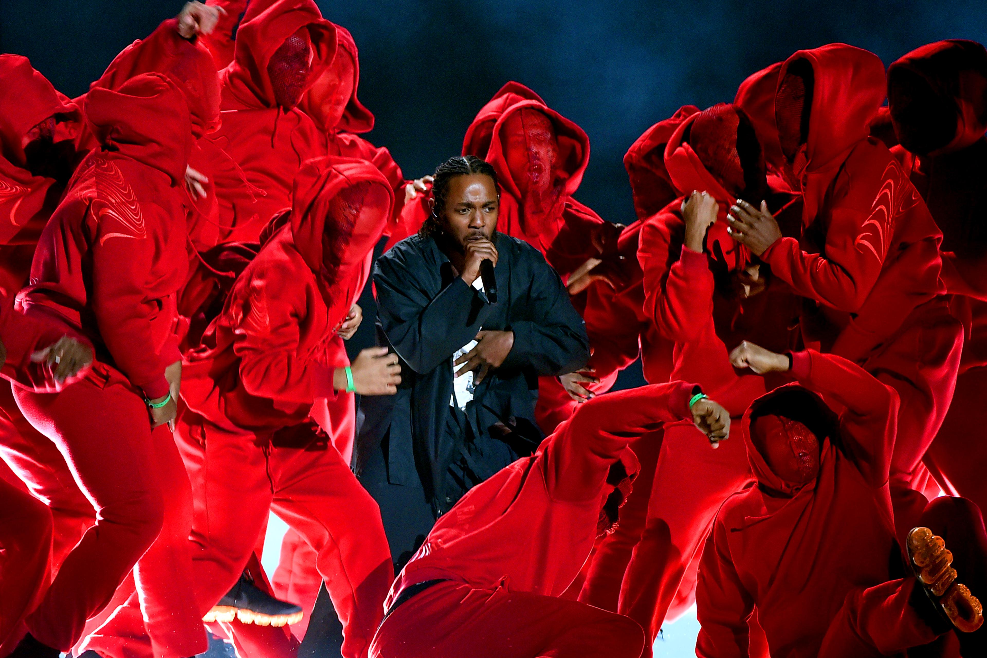 Photo of a performer singing surrounded by a group of dancers in red hooded outfits on stage under dramatic lighting.
