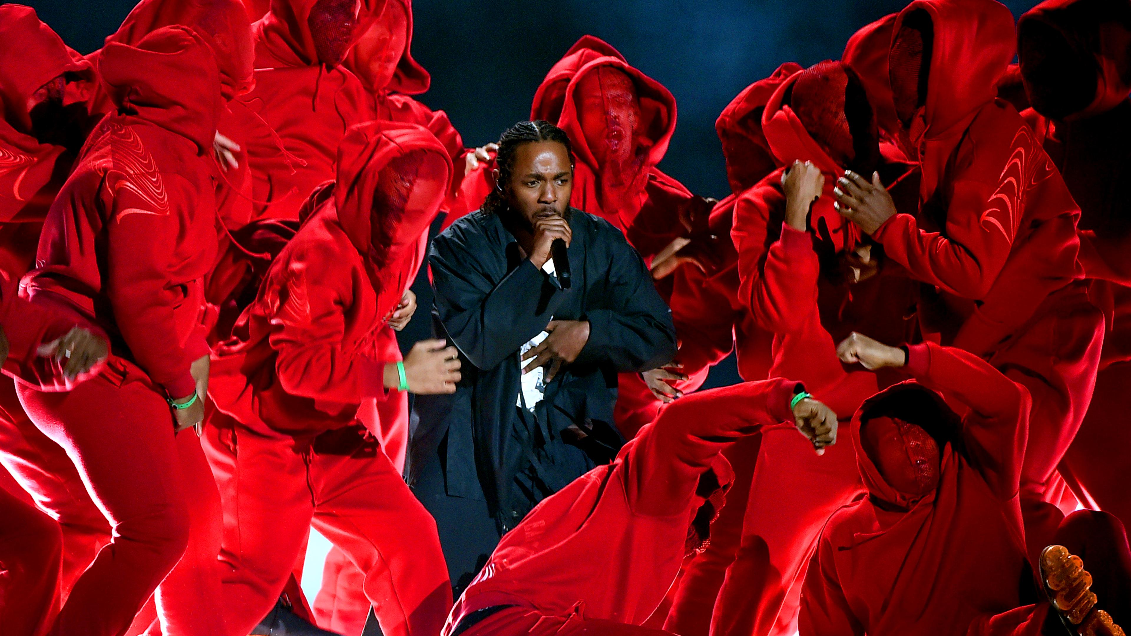 Photo of a performer singing surrounded by a group of dancers in red hooded outfits on stage under dramatic lighting.