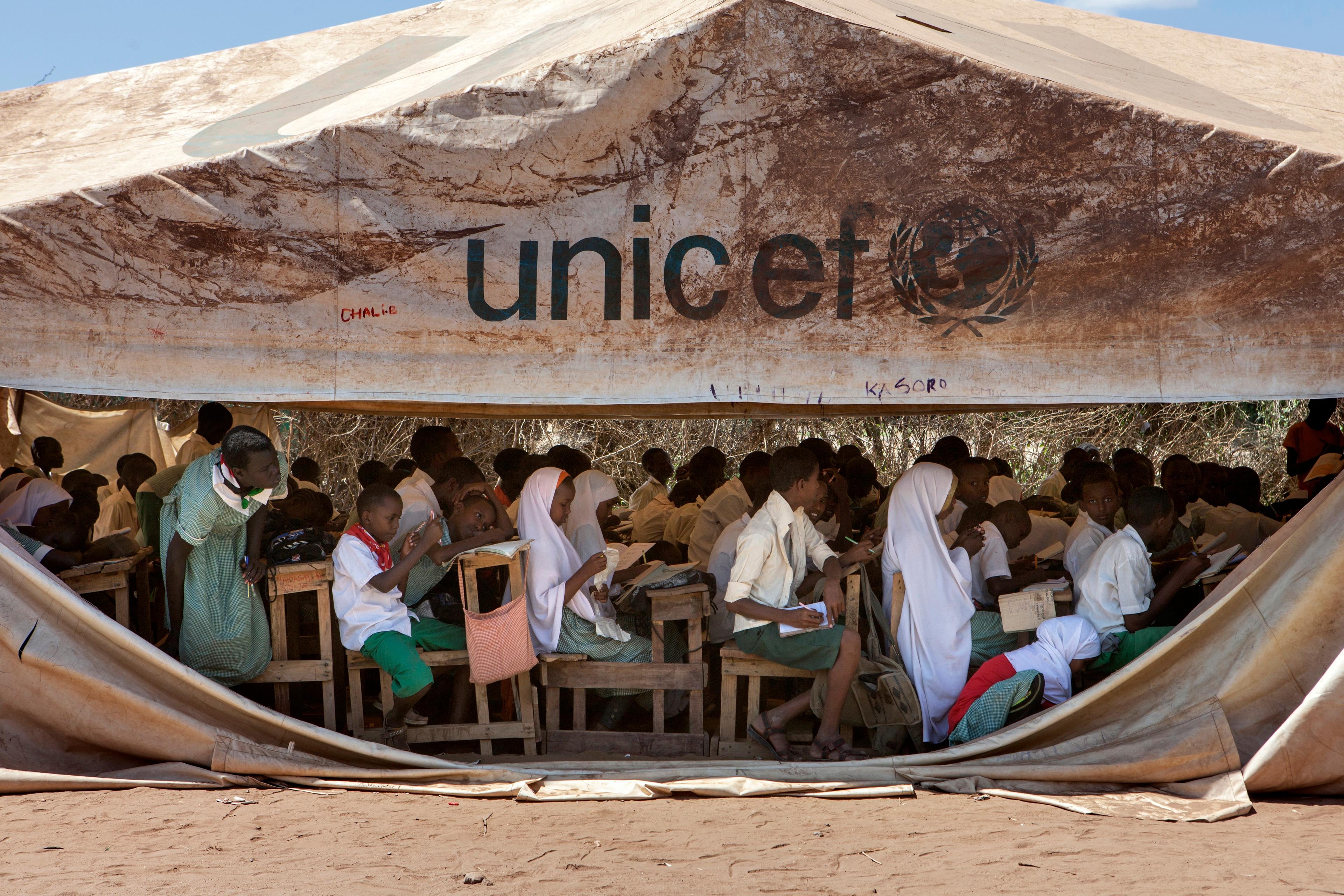 Photo of children in a UNICEF tent school, sitting at desks, studying outdoors in a humanitarian setting.