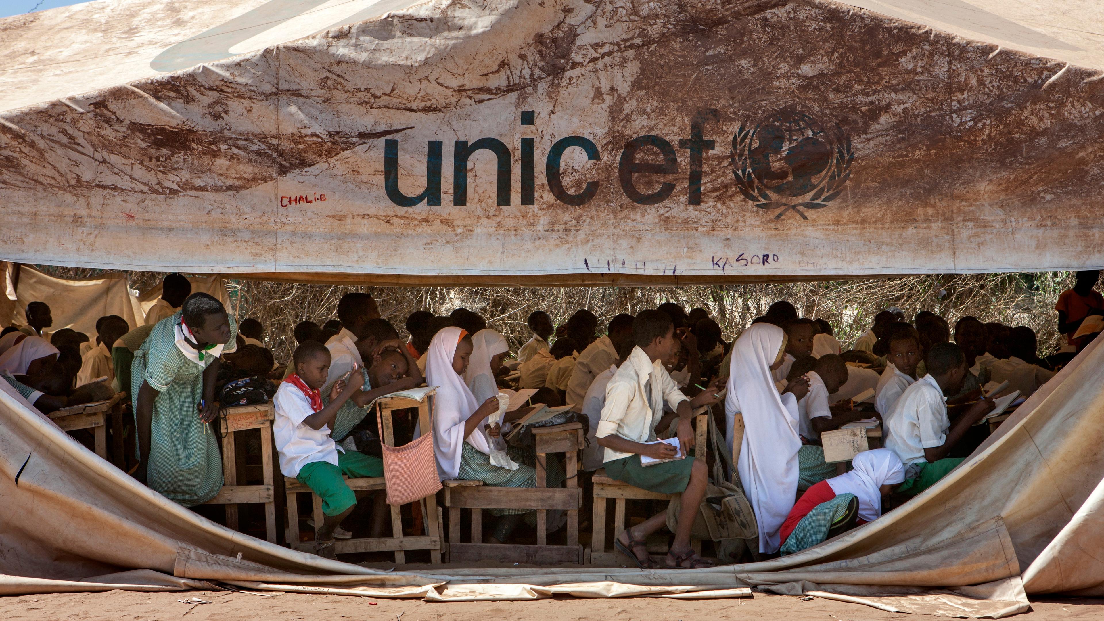 Photo of children in a UNICEF tent school, sitting at desks, studying outdoors in a humanitarian setting.