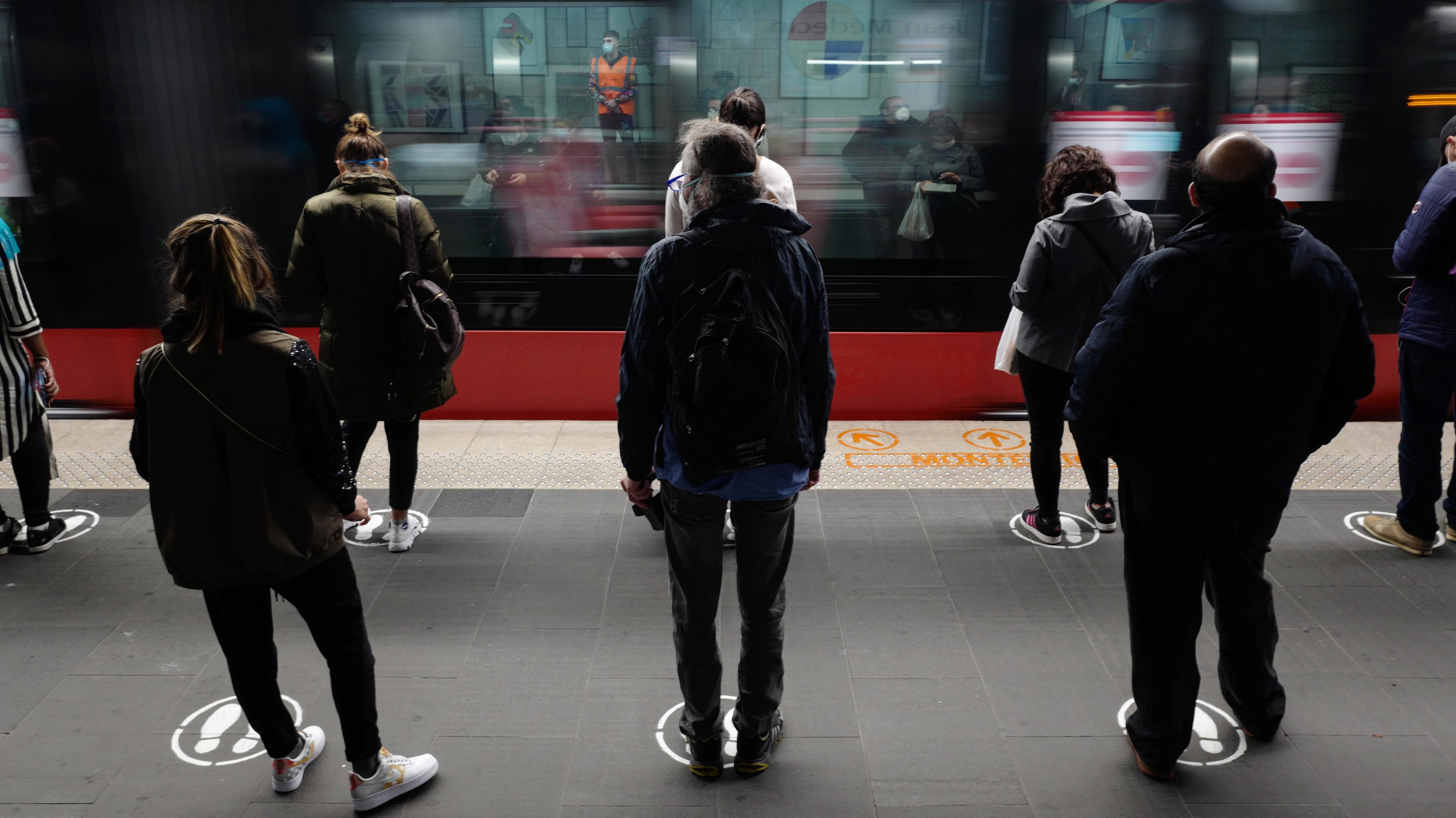 Photo of people waiting on a train platform standing on marked spots as a train speeds past in the background.