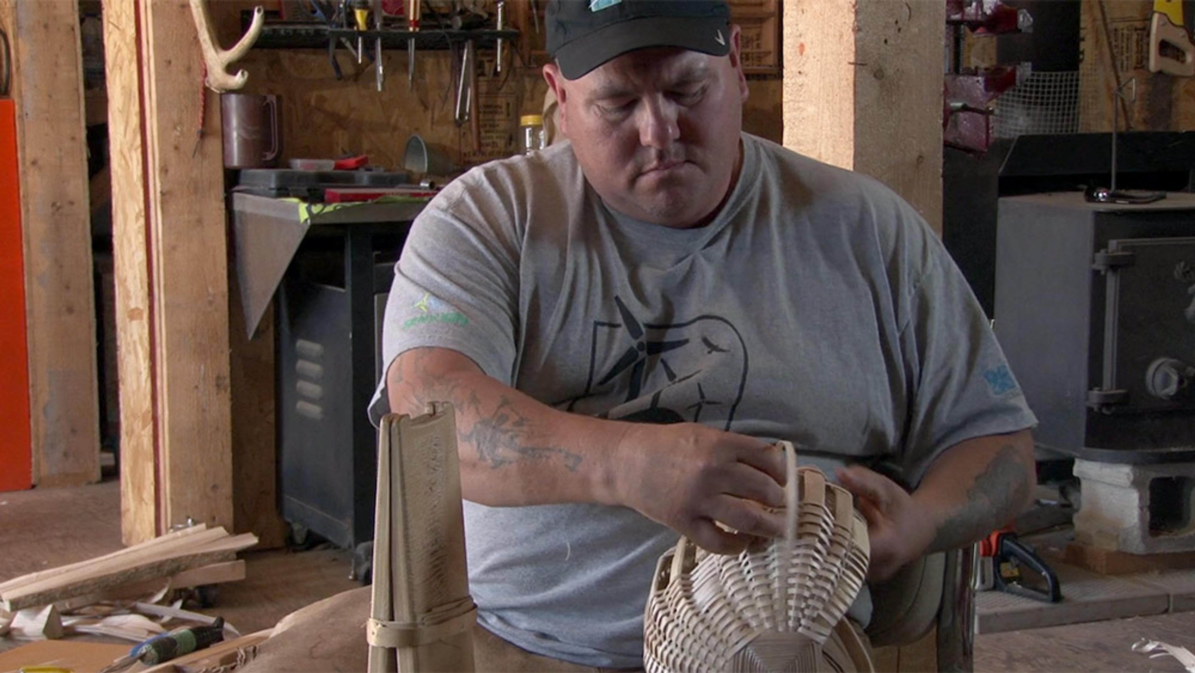 A large man with a tattoo on his arm, wearing a cap and T-shirt, making a basket in a workshop filled with tools, wooden beams and a wood-burning stove.