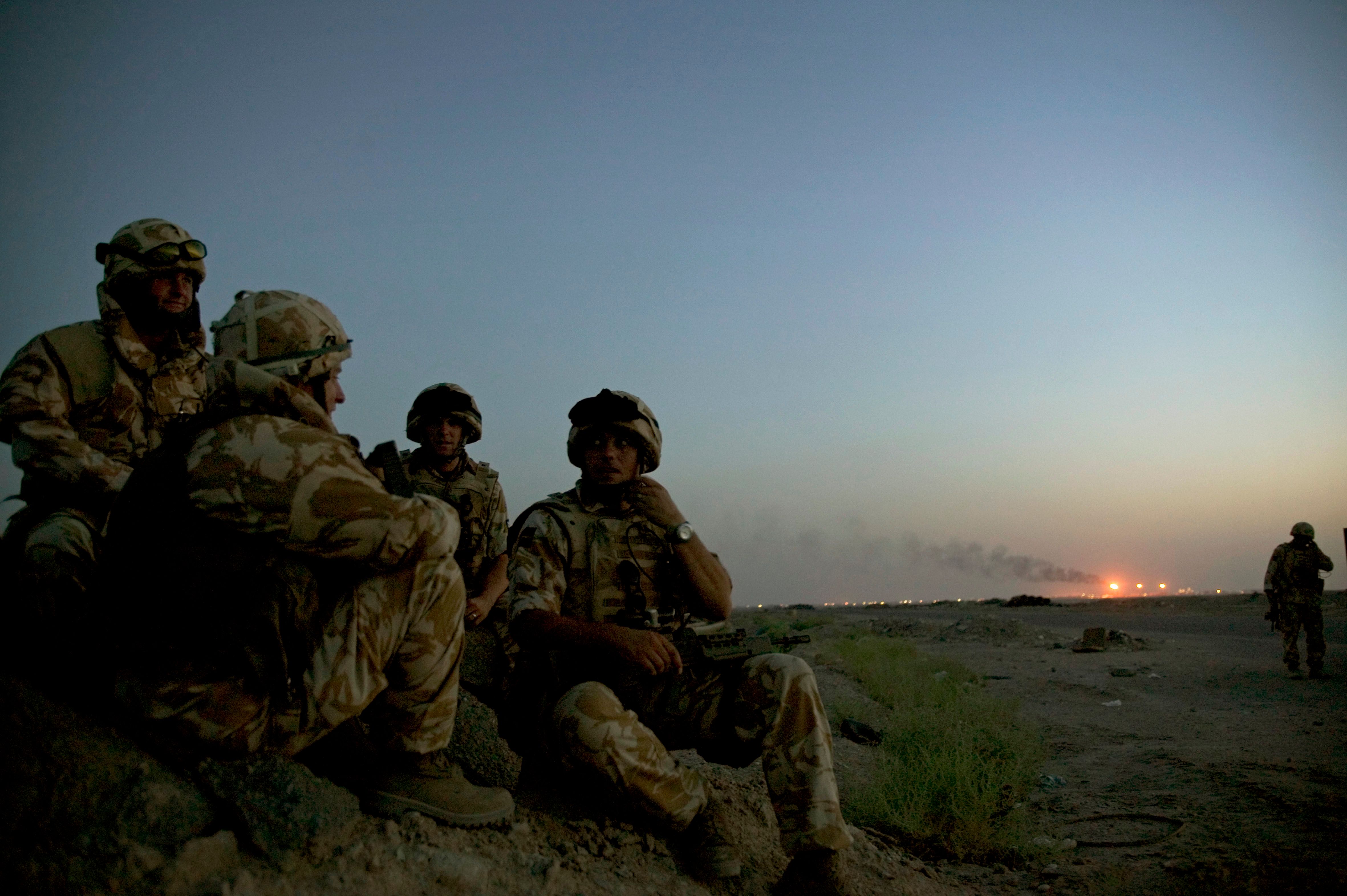 Photo of soldiers in camouflage sitting on a rocky terrain at dusk with a smoky horizon and distant lights in the background.