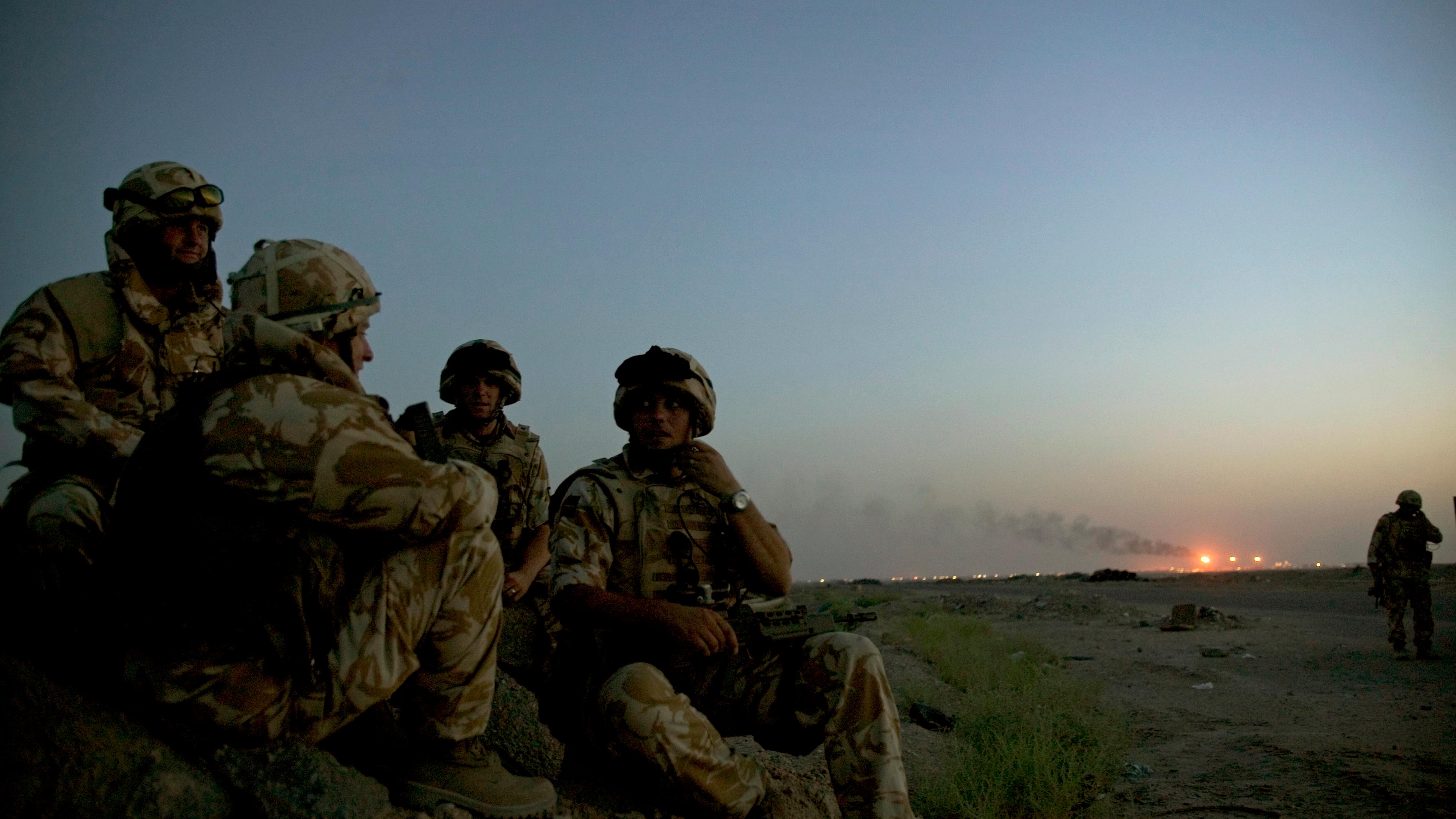 Photo of soldiers in camouflage sitting on a rocky terrain at dusk with a smoky horizon and distant lights in the background.