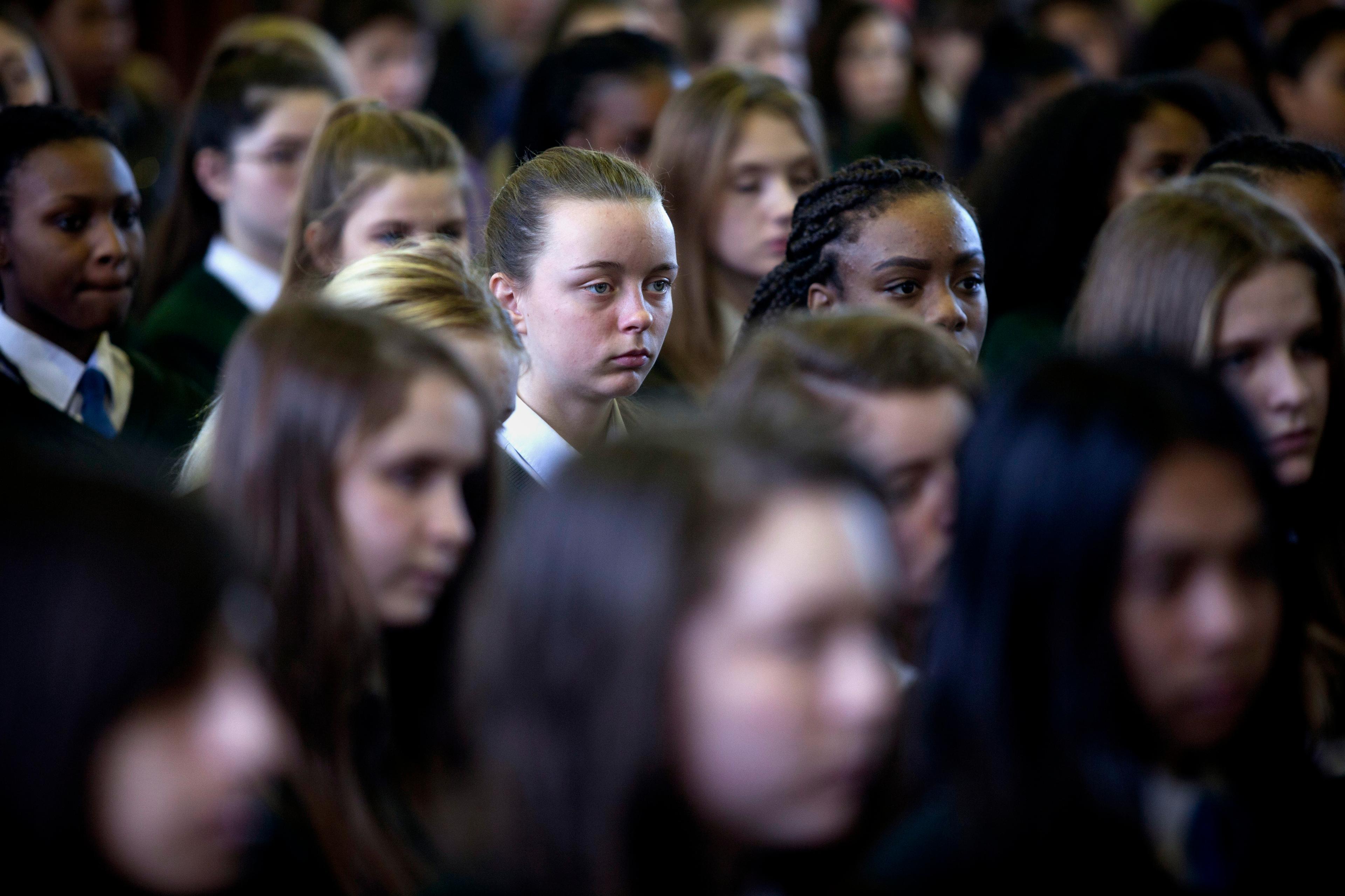 Photo of school assembly with pupils in uniform, many faces blurred. One student is in focus in the centre of the image.