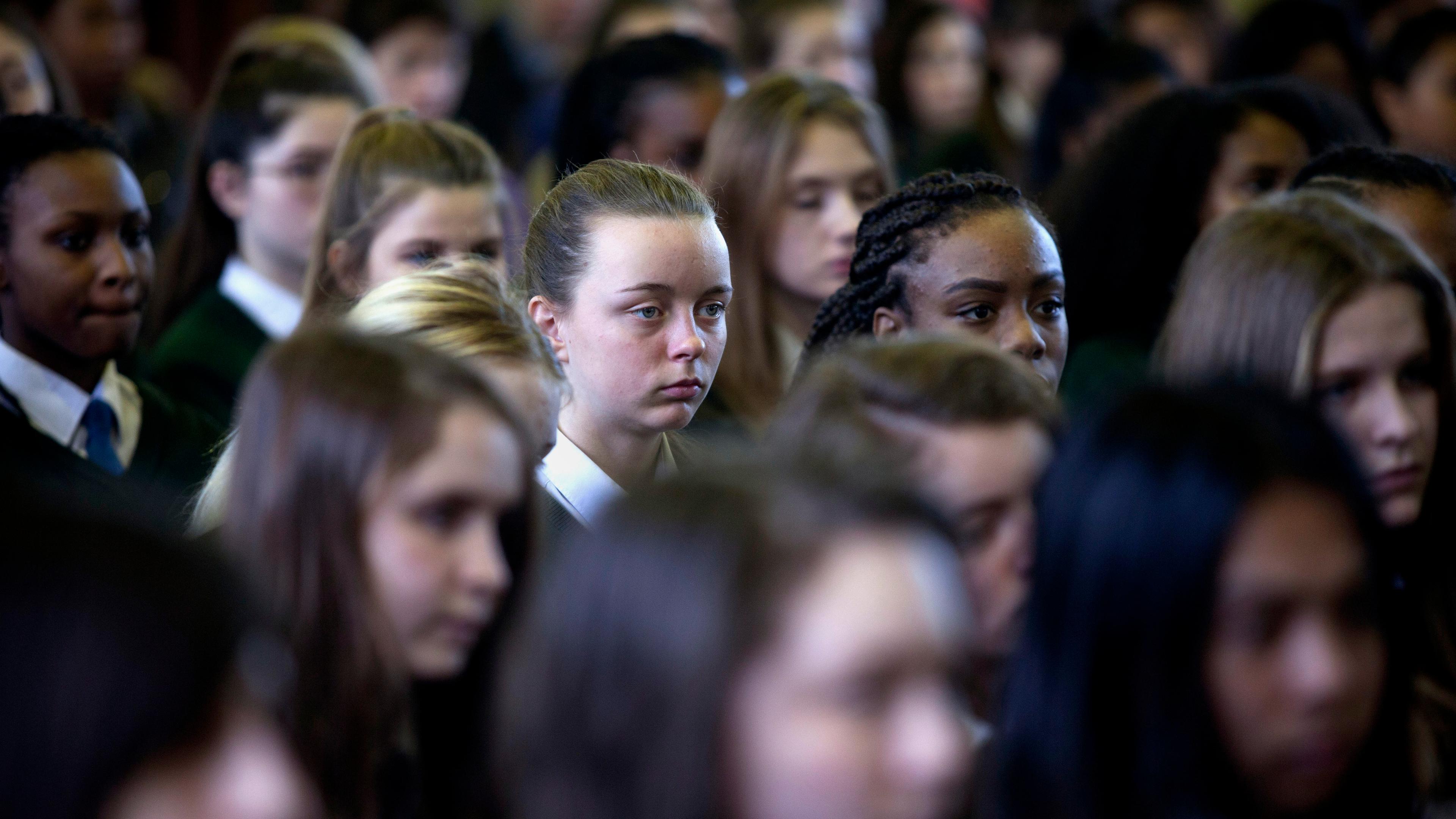 Photo of school assembly with pupils in uniform, many faces blurred. One student is in focus in the centre of the image.