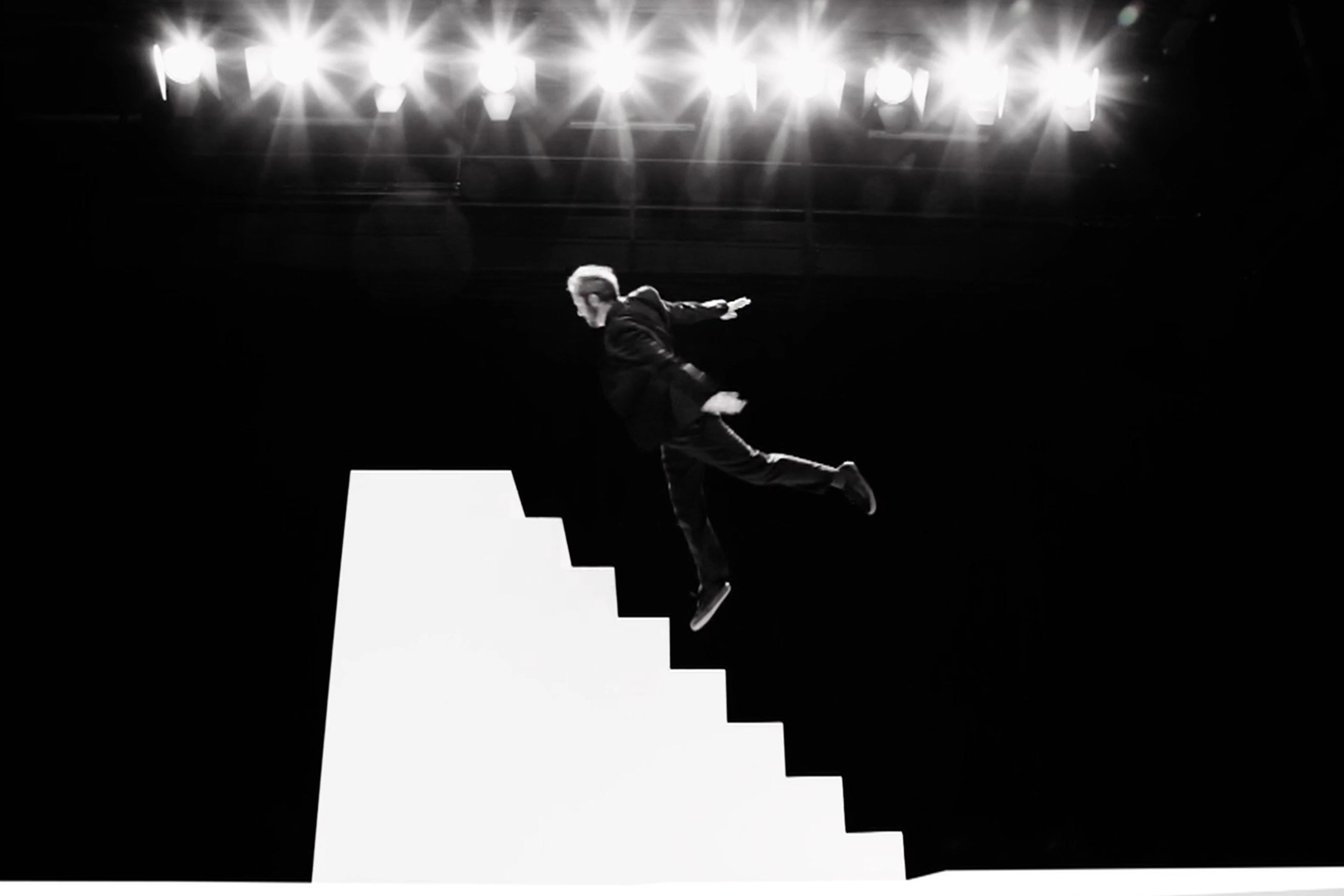 A film still of a man in a black suit airborne above a white staircase under bright stage lights against a dark background.