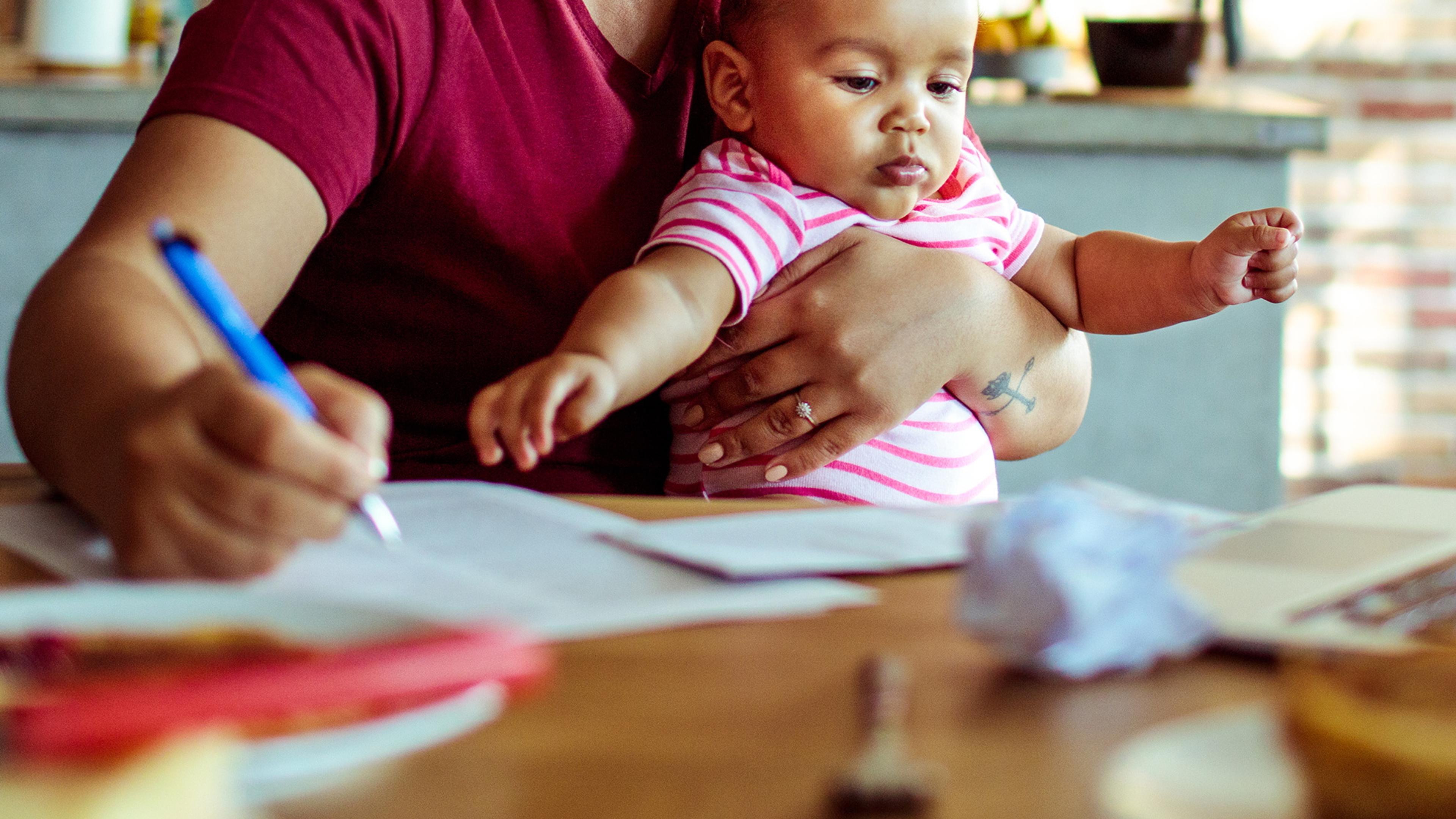 Photo of a person holding a baby while writing at a table with papers and a laptop.