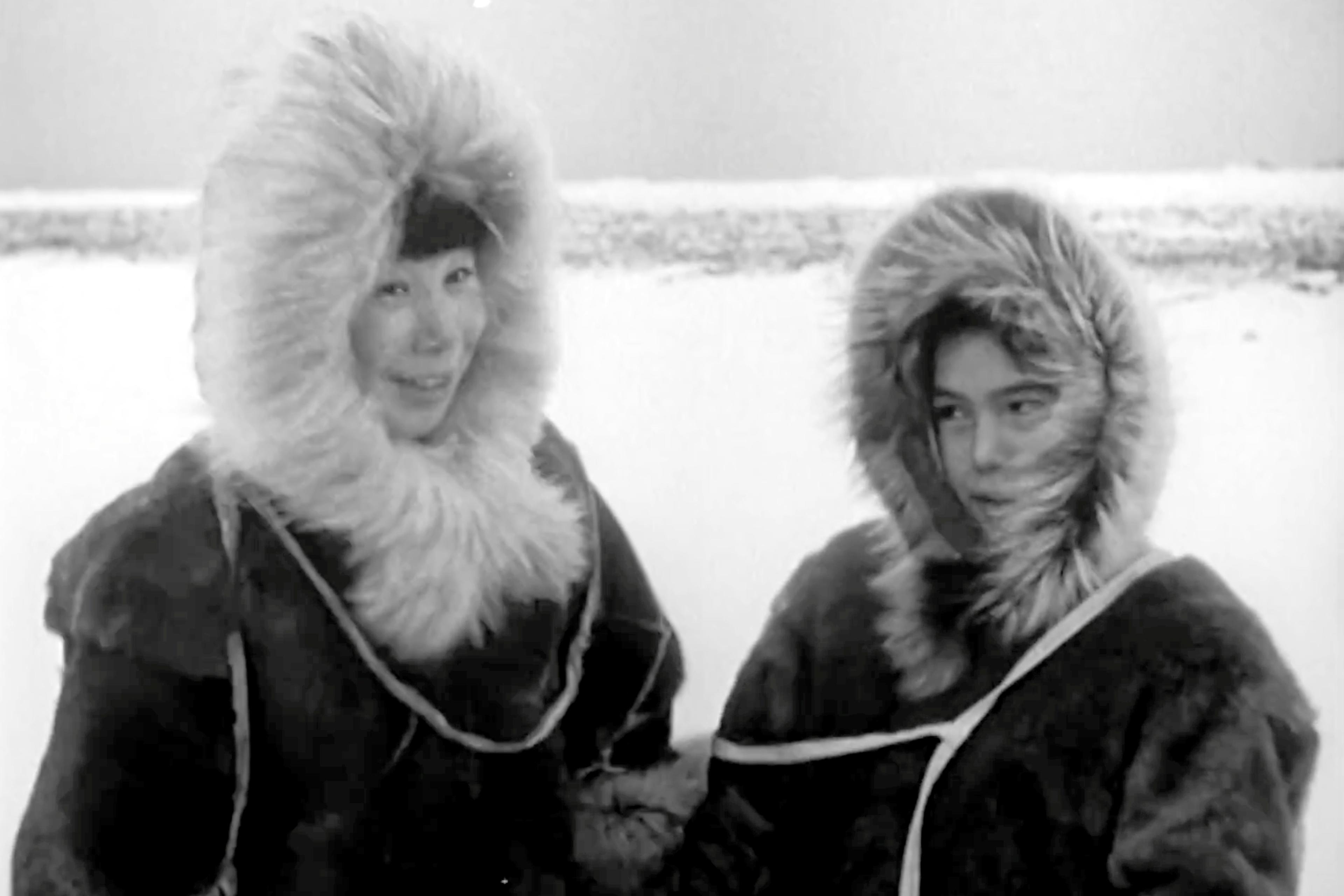 Black and white film still of two people in fur-lined parkas in a snowy landscape.