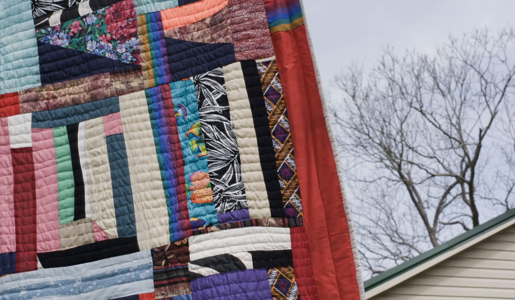 Colourful patchwork quilt hanging outside near leafless tree and house, showcasing various patterns and colours.