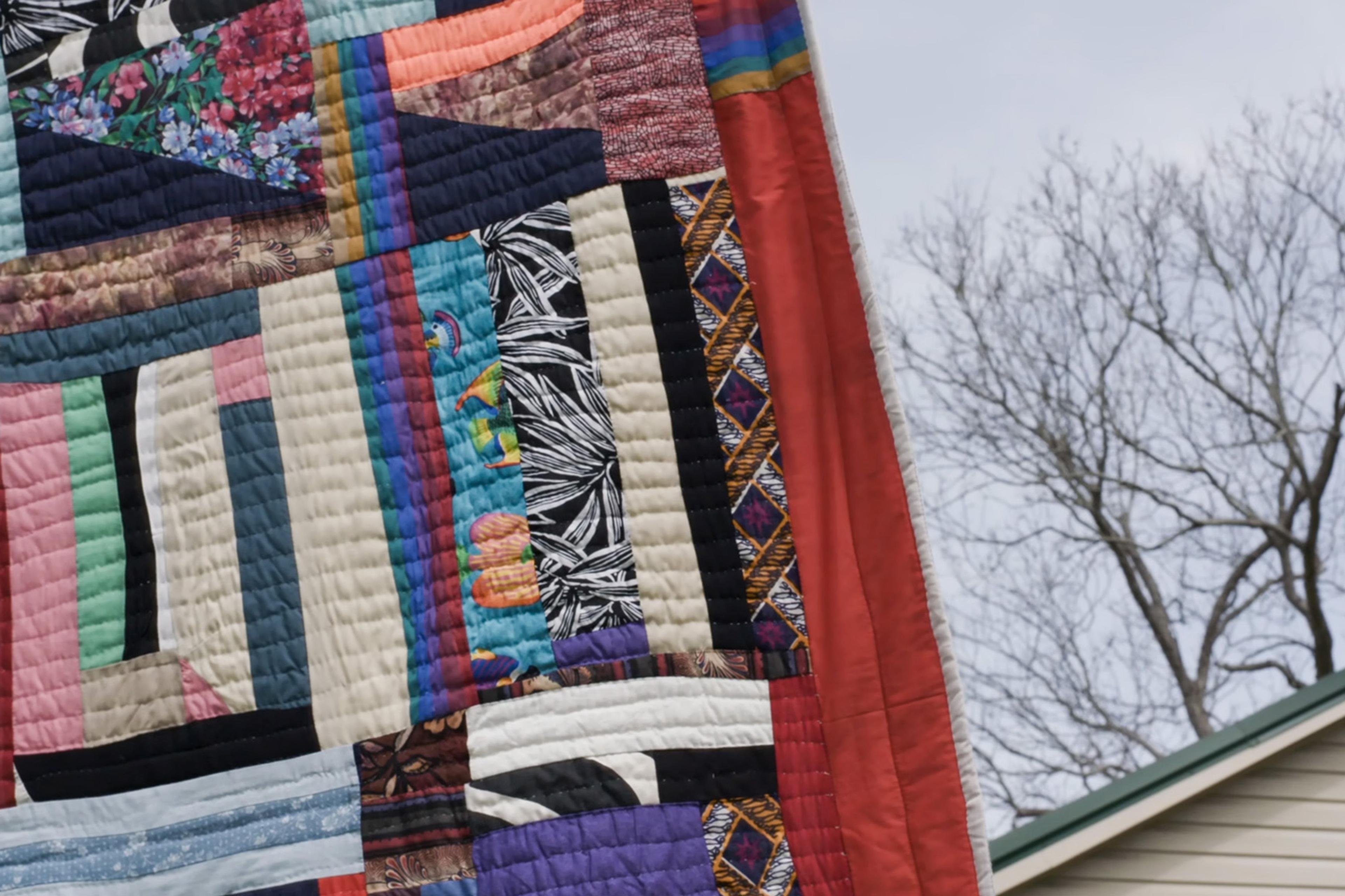Colourful patchwork quilt hanging outside near leafless tree and house, showcasing various patterns and colours.