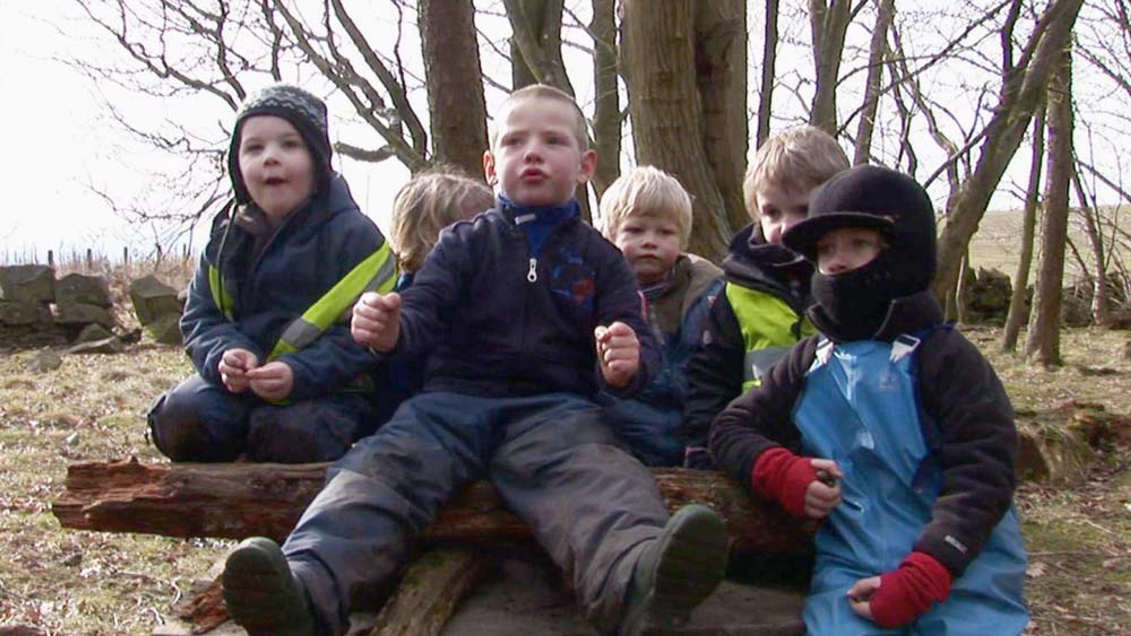 A group of children in winter clothing sitting on a large log outdoors in a forested area with bare trees in the background.