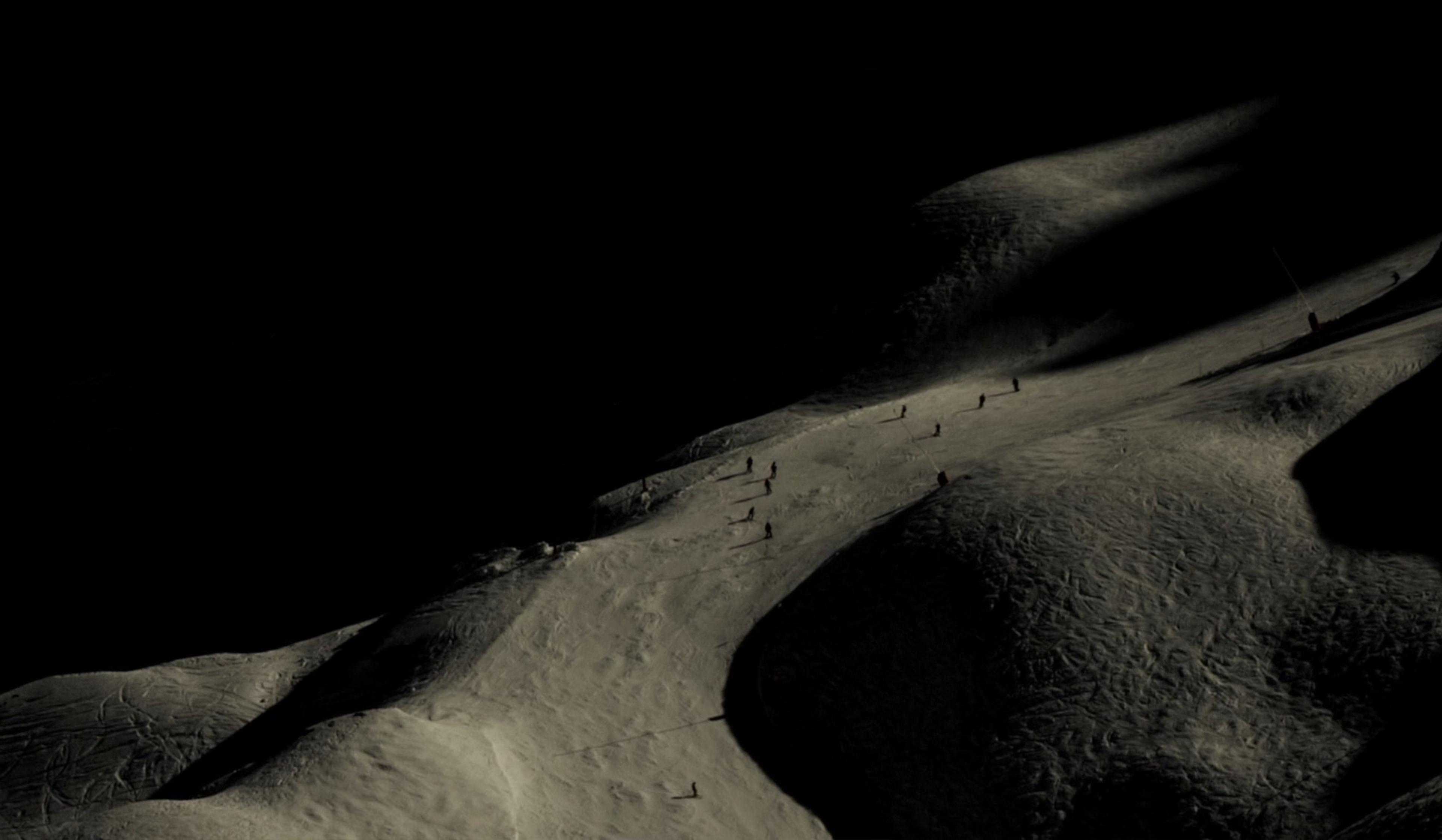 A dramatic photo of a snowy mountain slope at night with scattered skiers casting long shadows on the illuminated path.