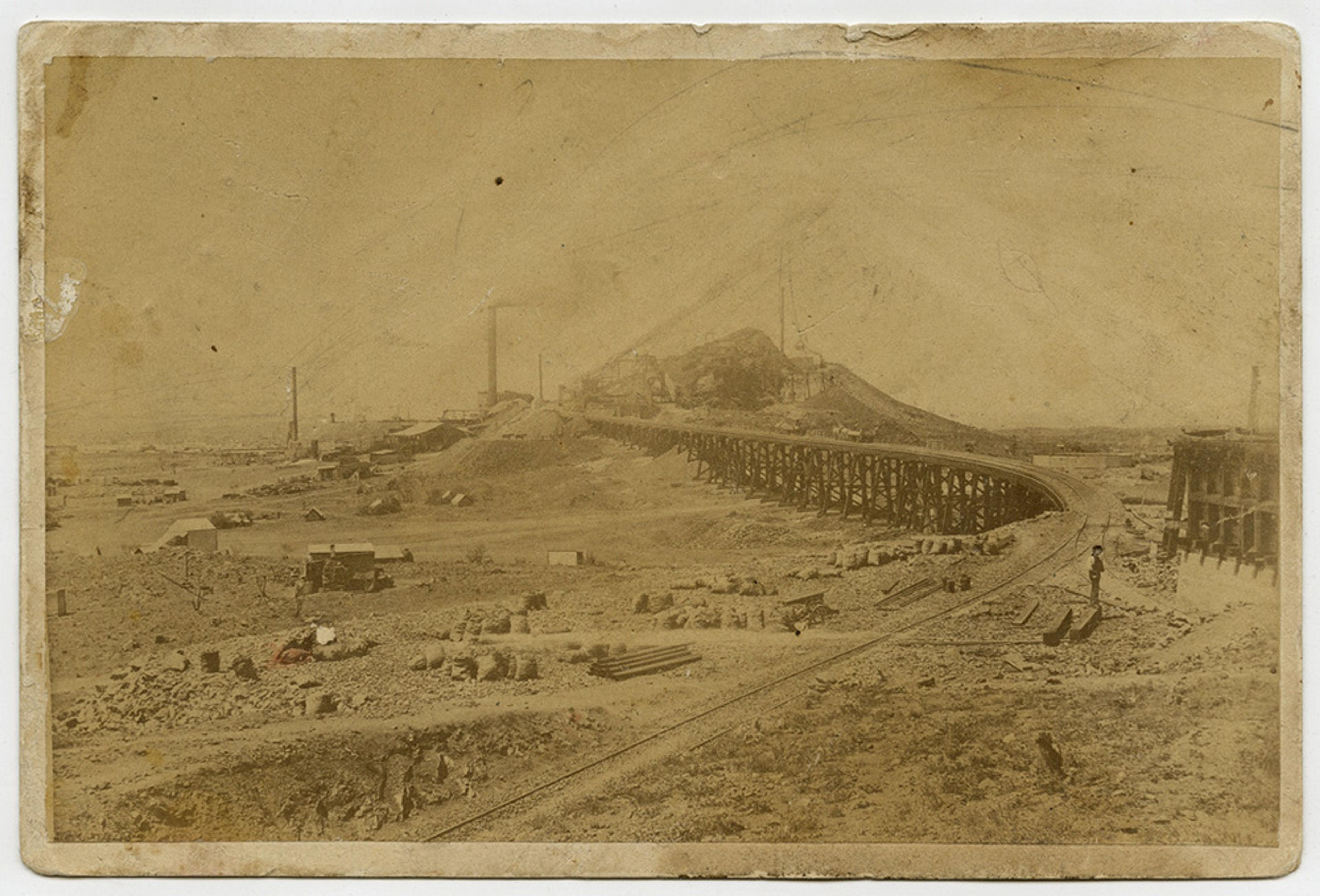Vintage sepia photo of an industrial landscape featuring a railway, large piles of materials, and distant buildings with smoke stacks.