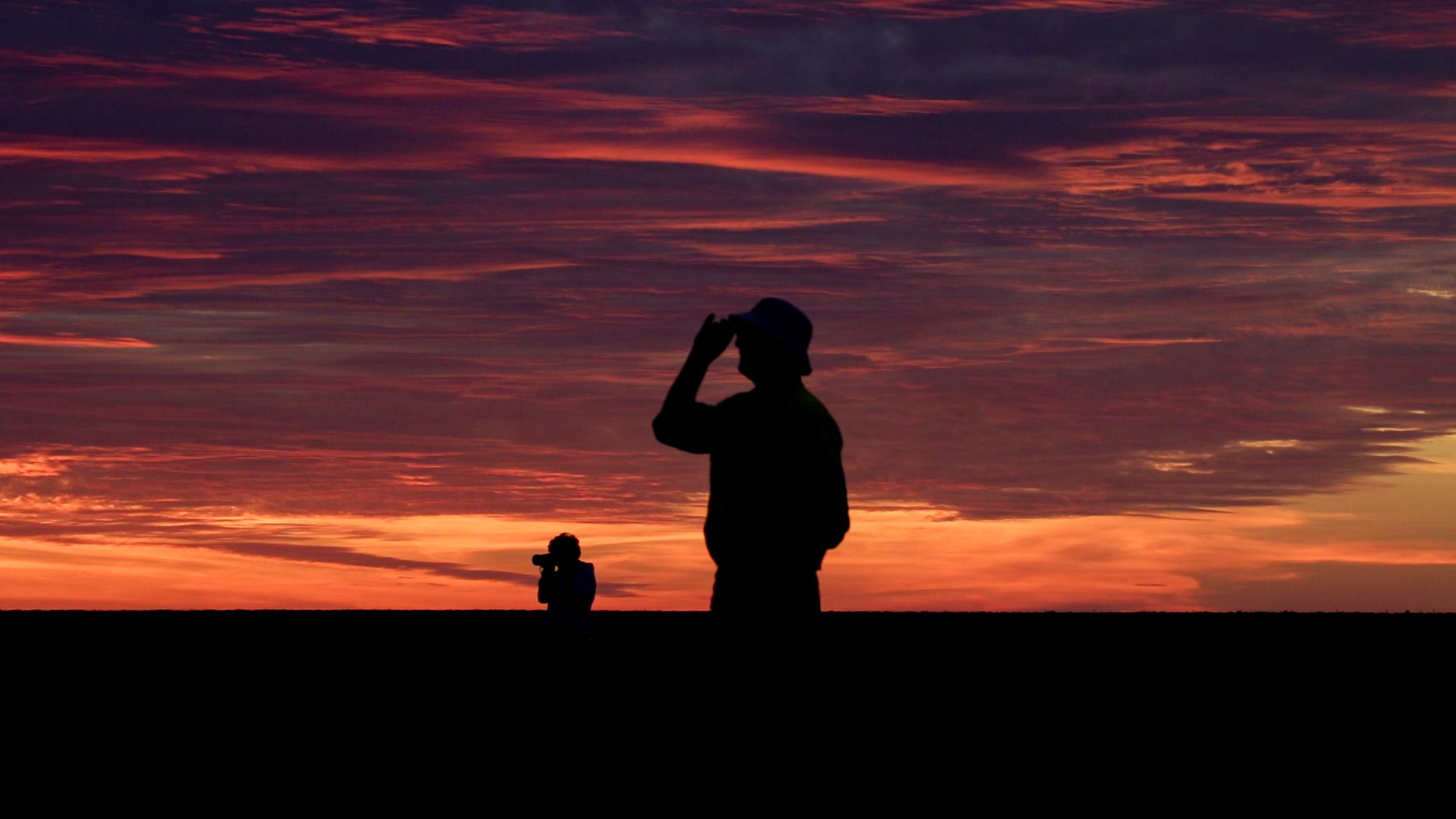 Two silhouetted people stand against a dramatic orange and purple sunset sky.